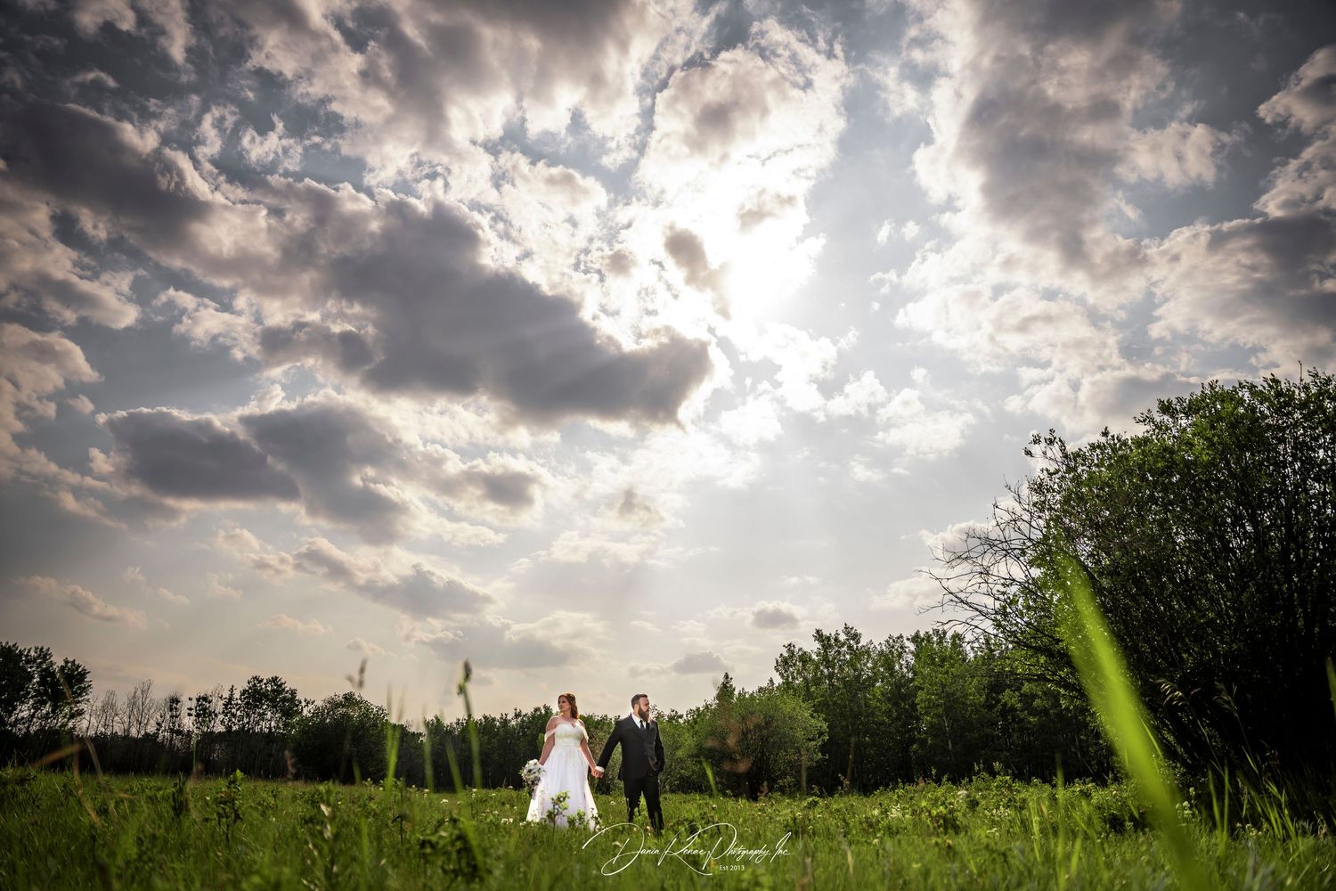 Bride and groom walking through lush green field under dramatic cloudy sky on their wedding day.
