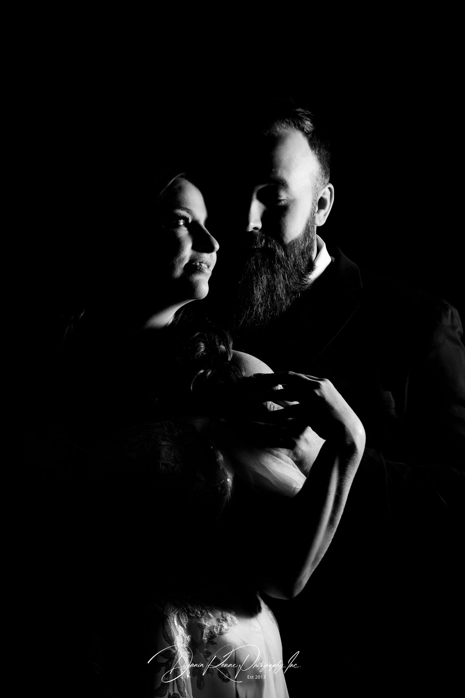 Black and white portrait of a bearded man embracing a woman holding a newborn baby against dark background.