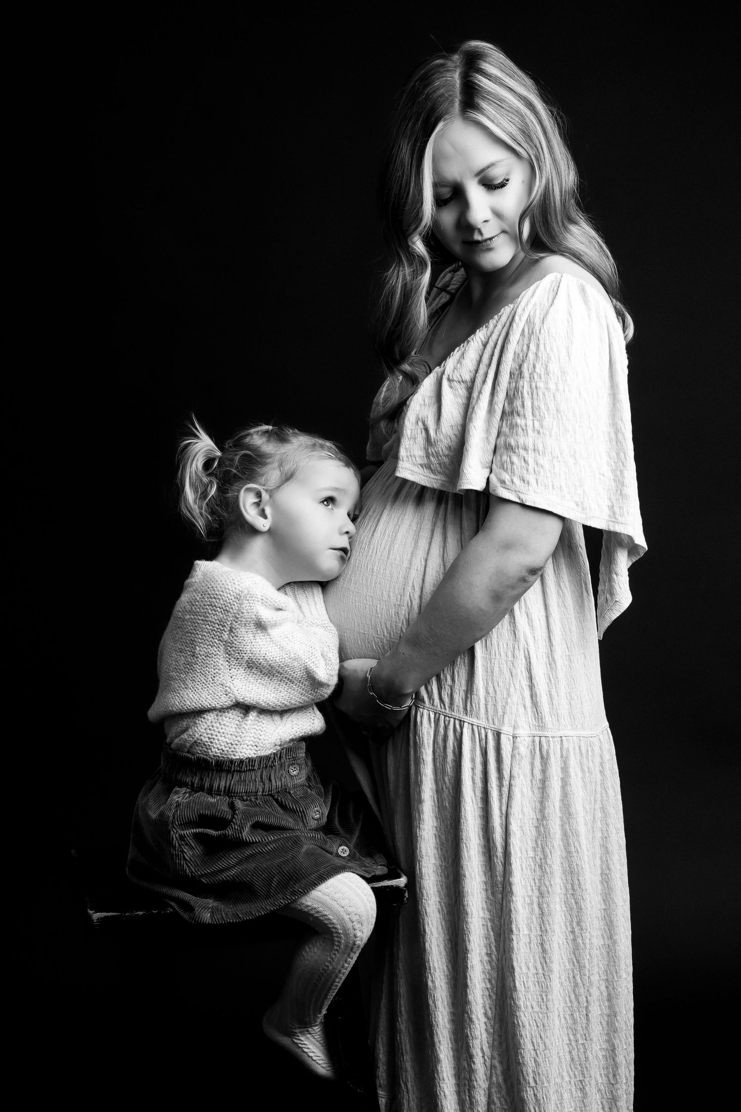 Black and white maternity photo of pregnant woman in dress with toddler girl touching her baby bump.