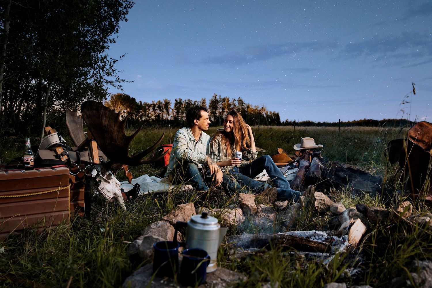 Friends gathered around a campfire at dusk, enjoying an outdoor camping trip in a scenic field.