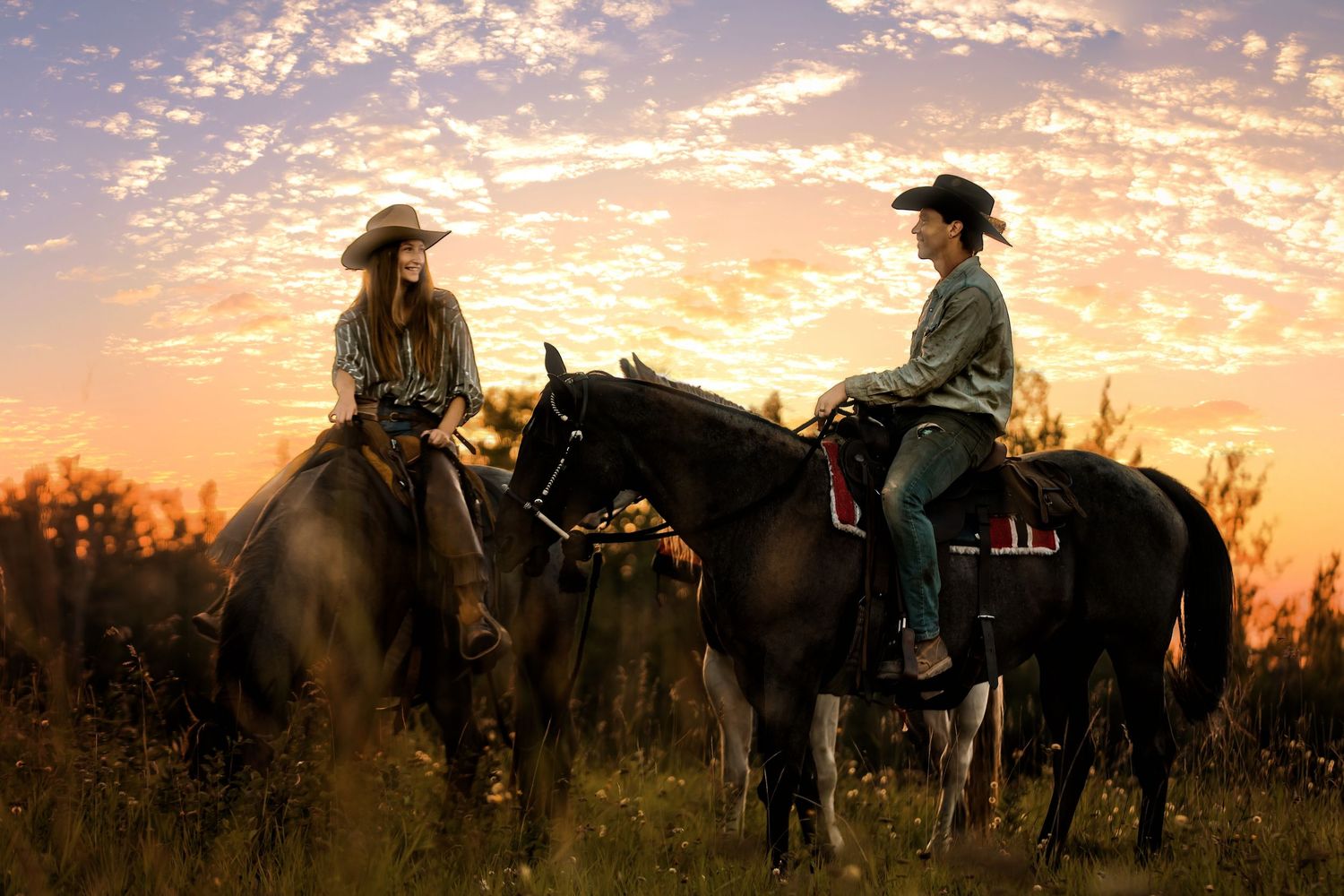 Two cowboys on horseback at golden sunset, wearing hats in a scenic field.