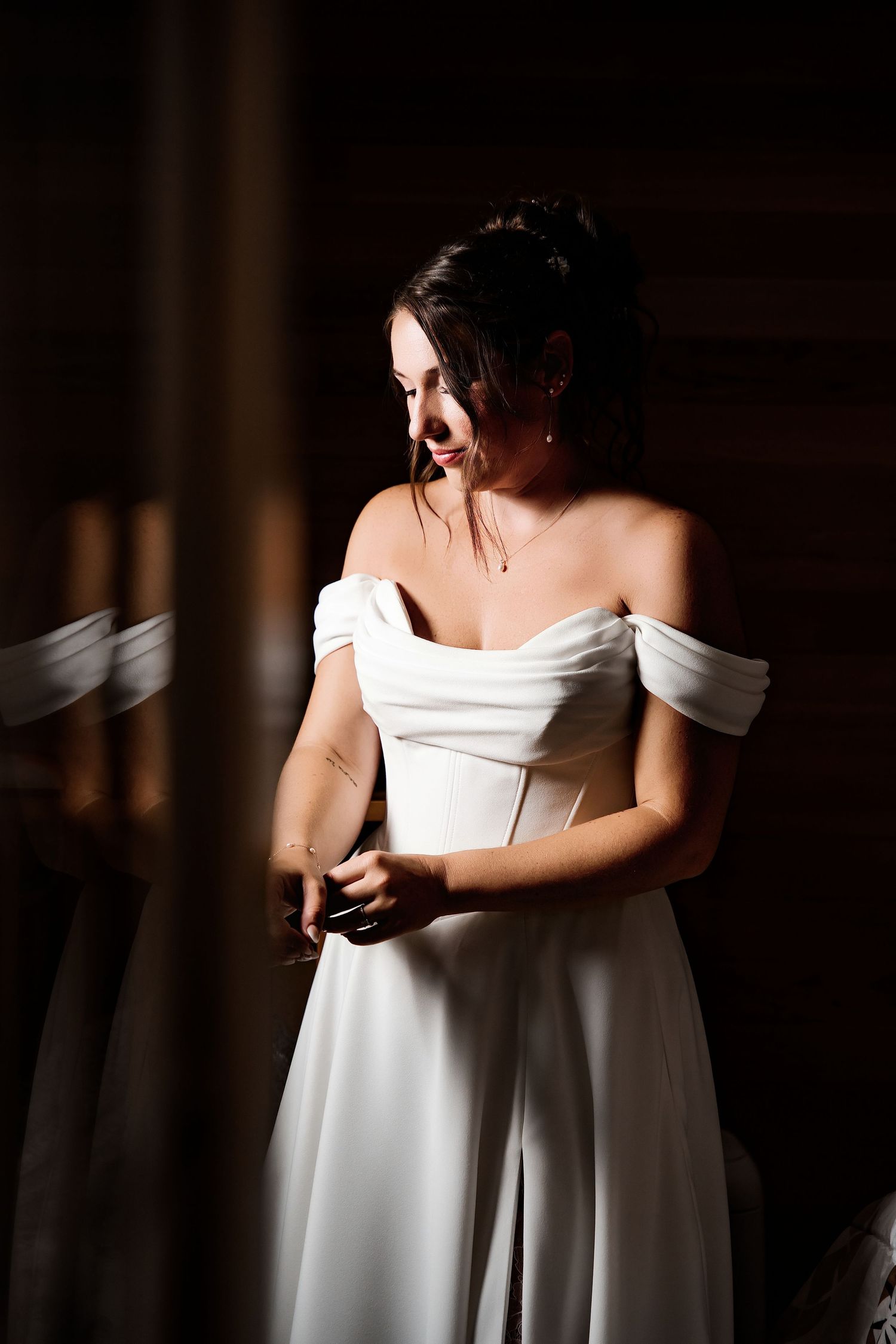Bride in elegant off-shoulder white wedding gown standing in dramatic lighting, looking down gracefully.