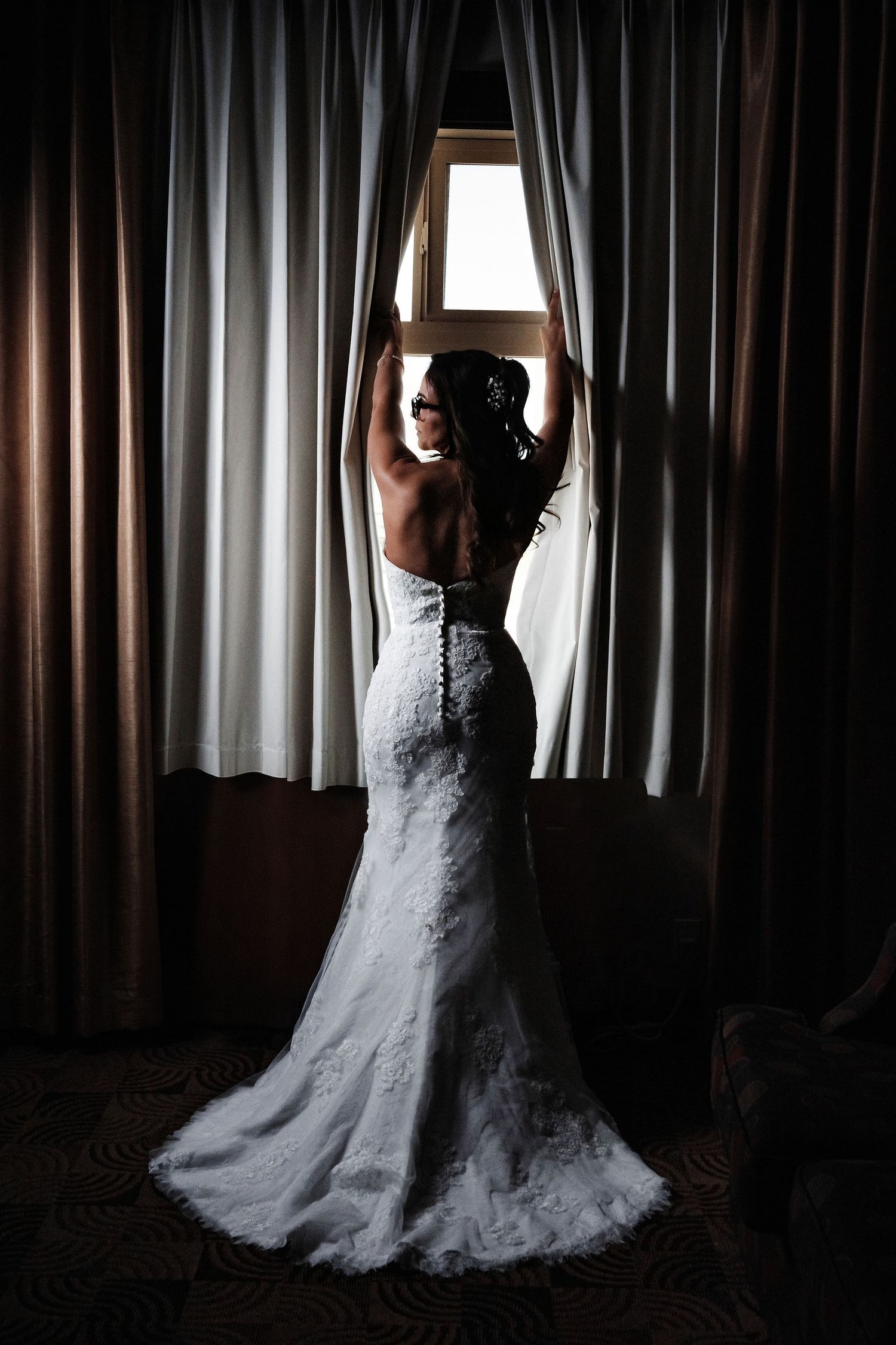 Bride in backless lace wedding gown stands before a curtained window, silhouetted in soft light.