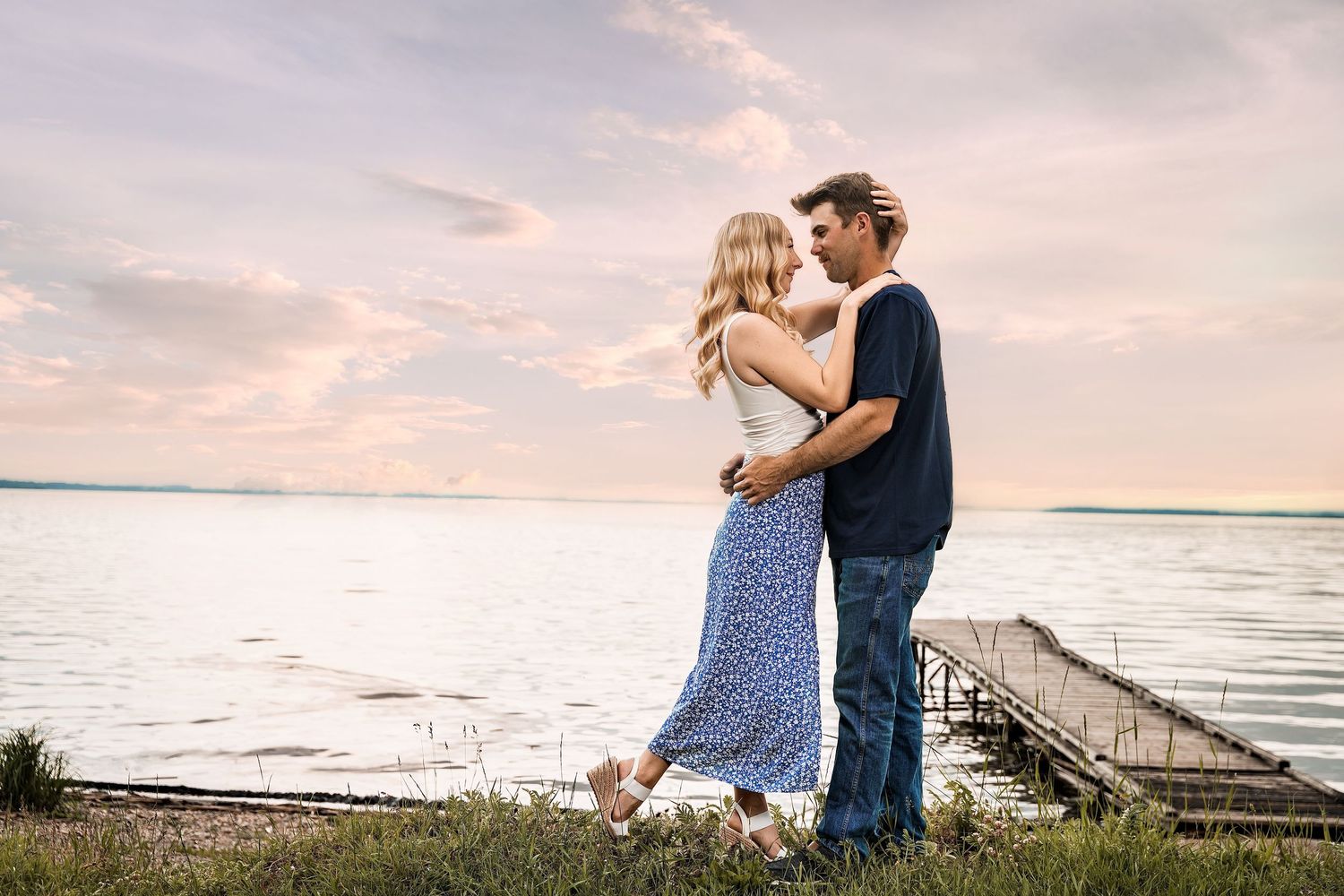 A romantic couple embraces near a lakeside dock at sunset, with a colorful sky and calm water in the background.