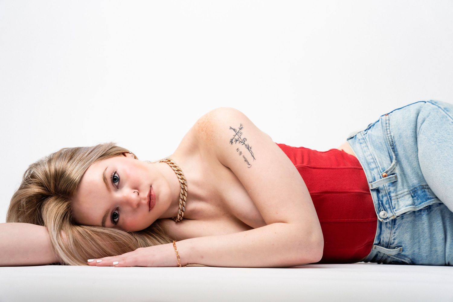 Young blonde woman lying down in red top and jeans, showing arm tattoo, posing against white background.