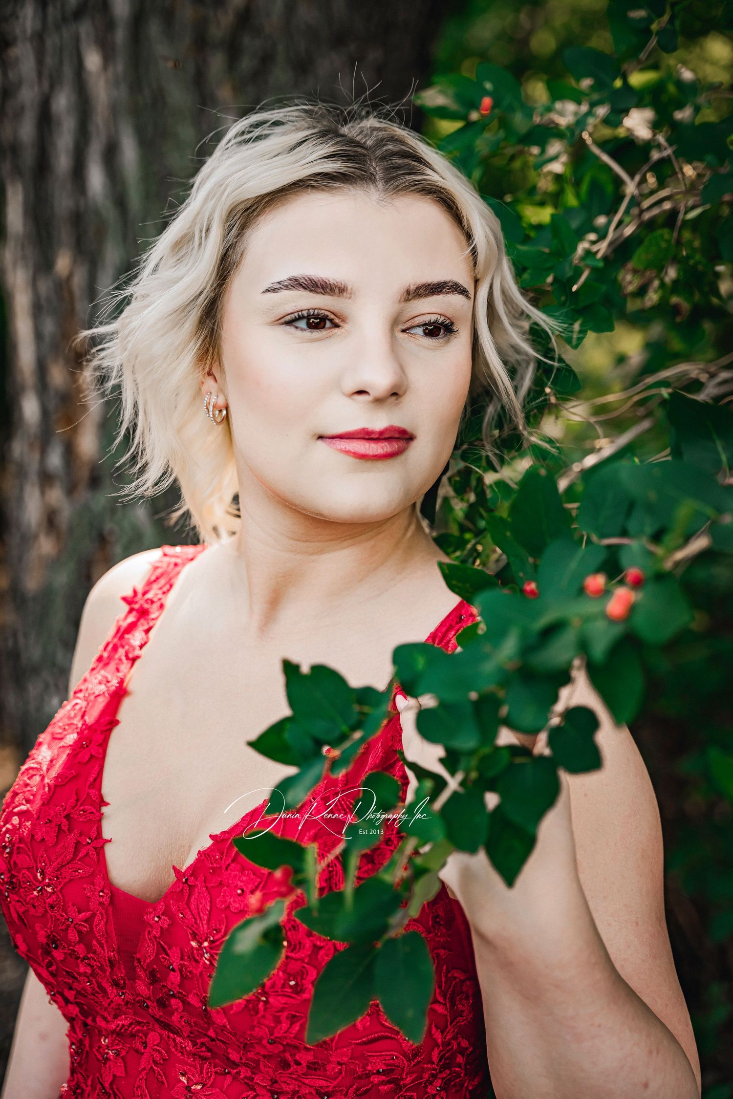 Young blonde woman in red lace dress posing outdoors near green foliage and red berries for a portrait photo.
