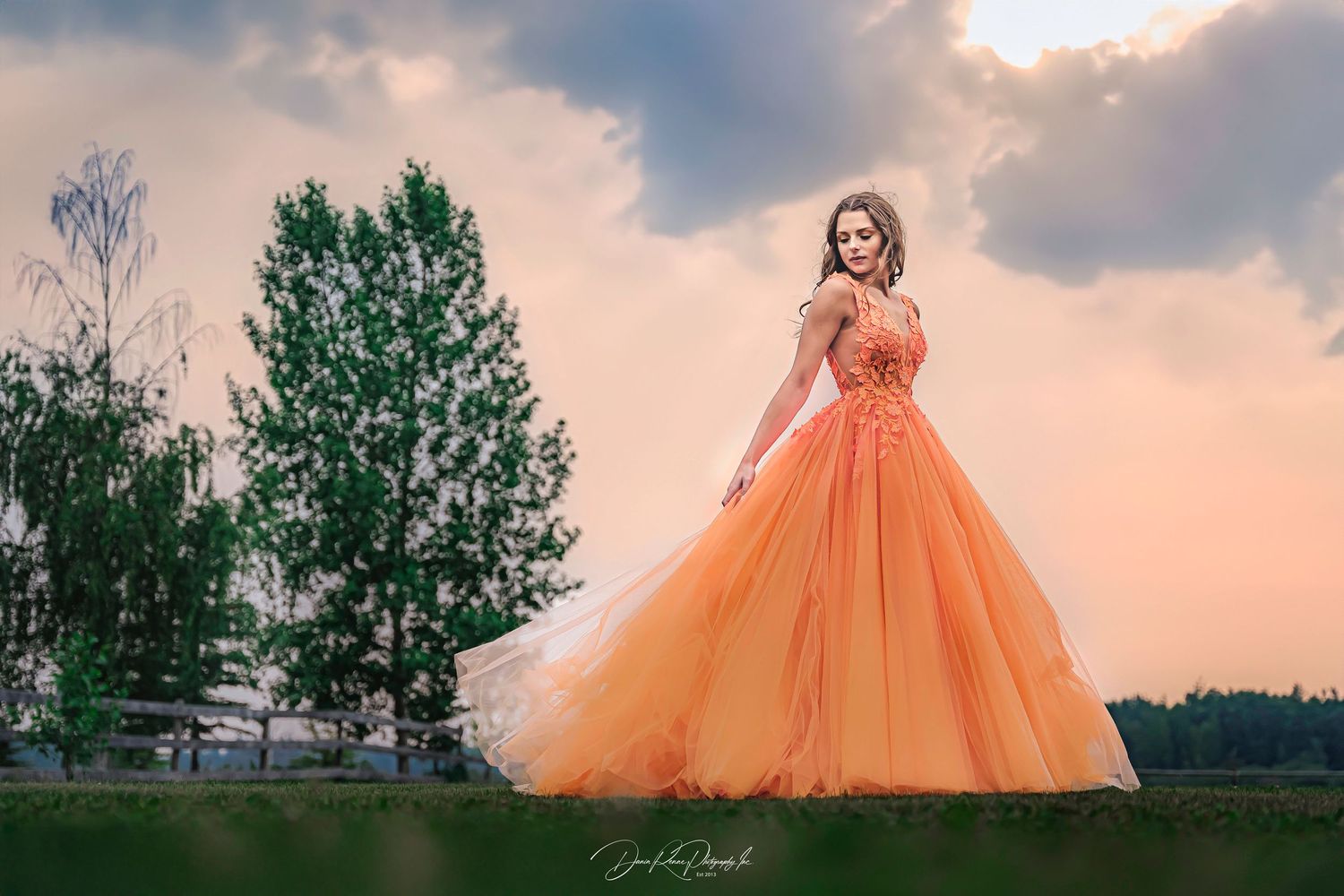 Young woman in elegant orange ball gown posing outdoors at sunset with lush green trees in background.