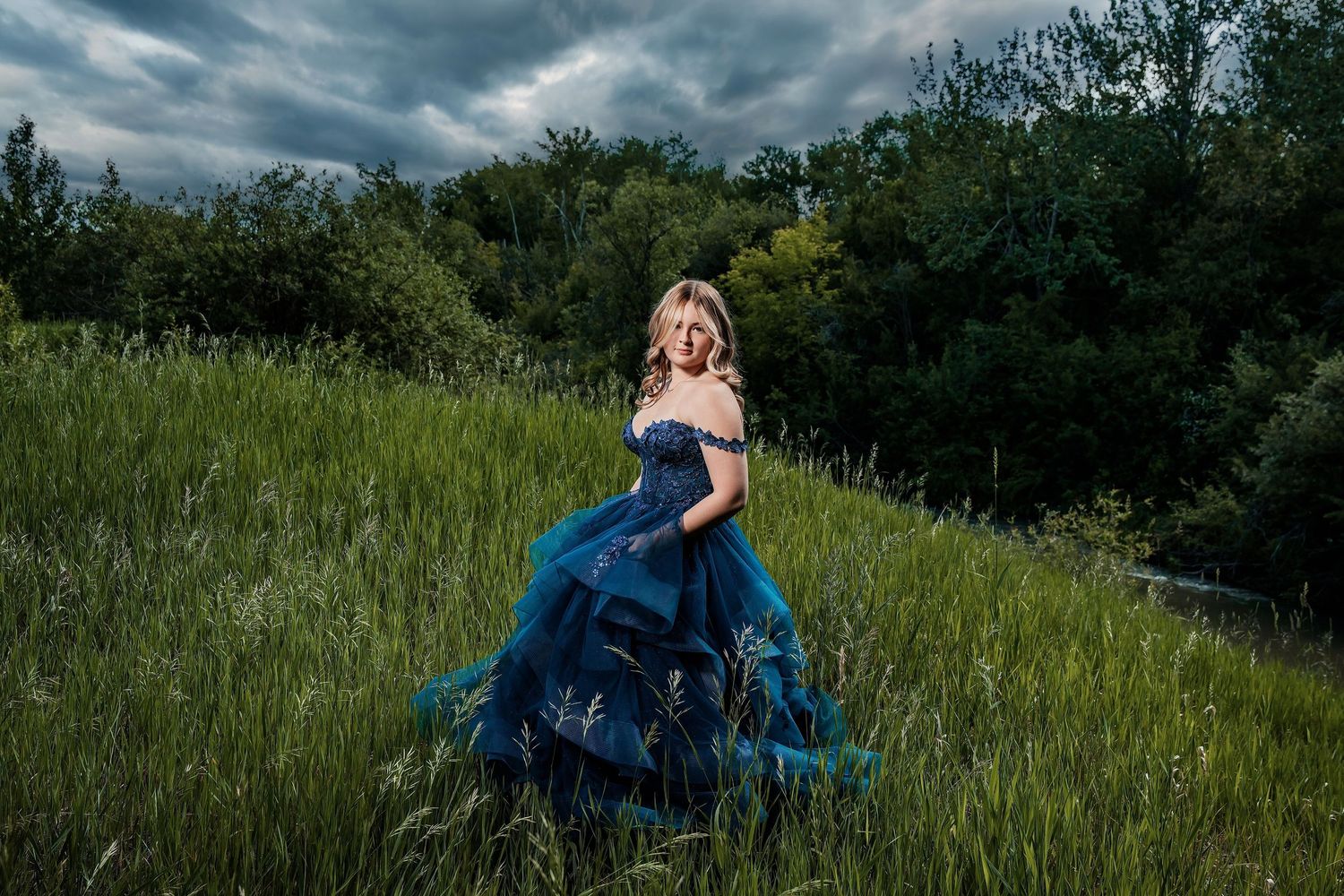 Woman in teal ball gown standing in lush green field with dramatic cloudy sky and trees in background.