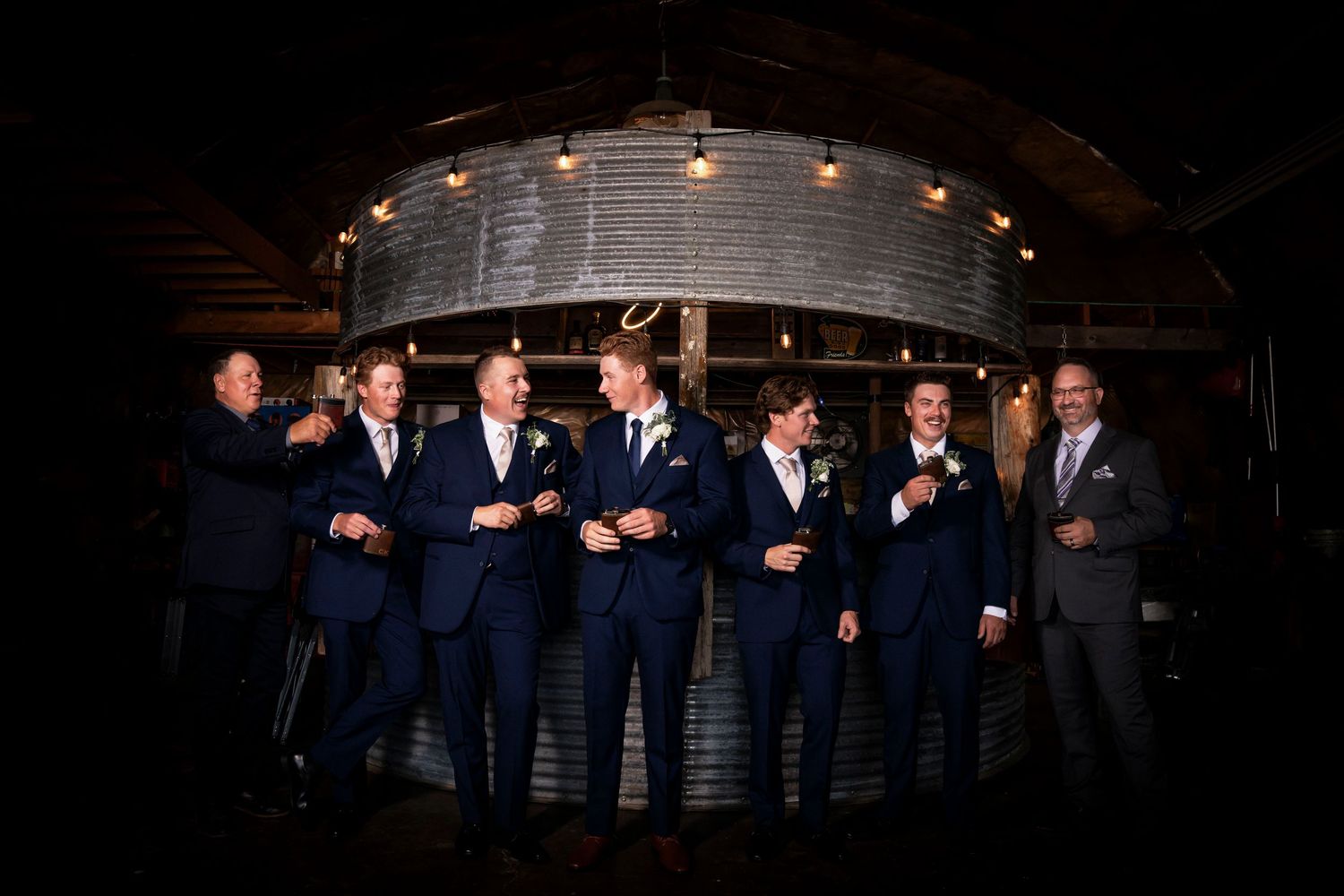Groomsmen in navy blue suits laughing and celebrating together in a rustic barn venue with warm string lights.