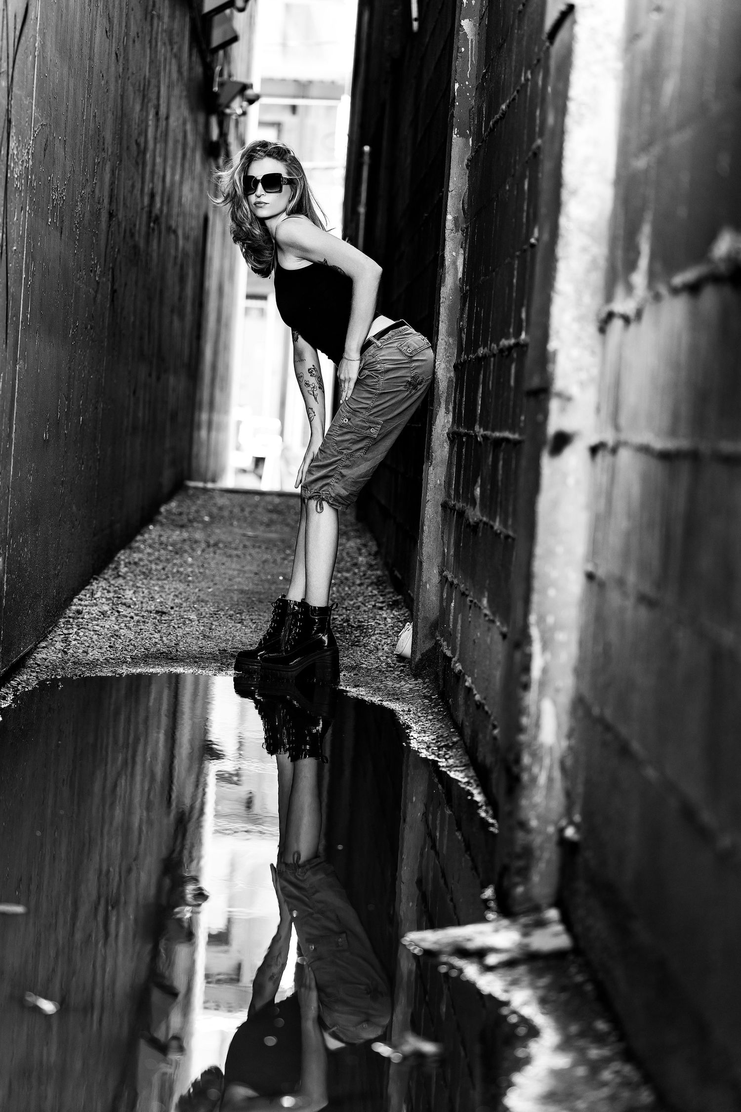 Black and white photo of a stylish woman in sunglasses posing in a narrow alley with a puddle reflection.
