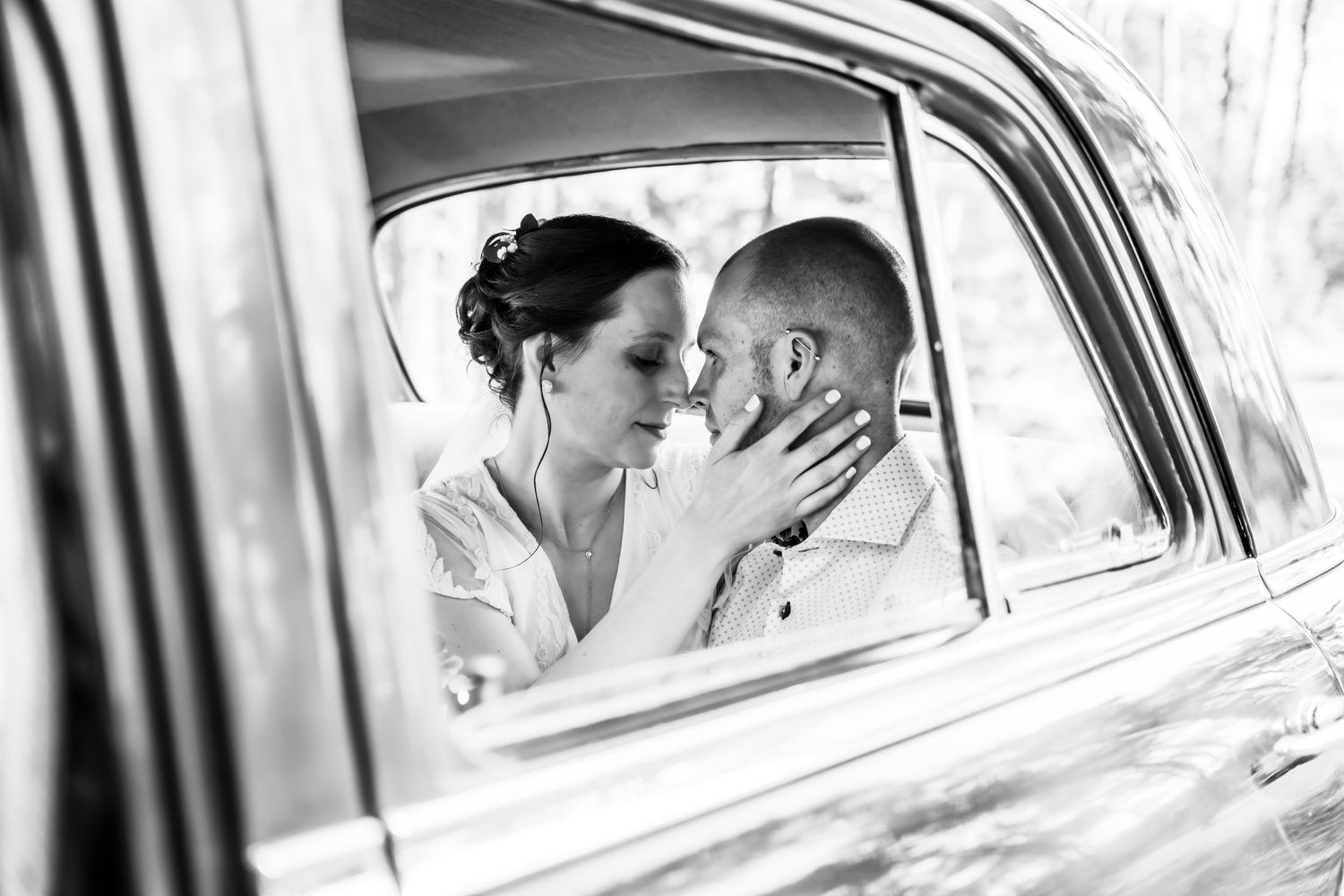 Black and white photo of a bride and groom sharing a romantic moment inside a vintage car on their wedding day.