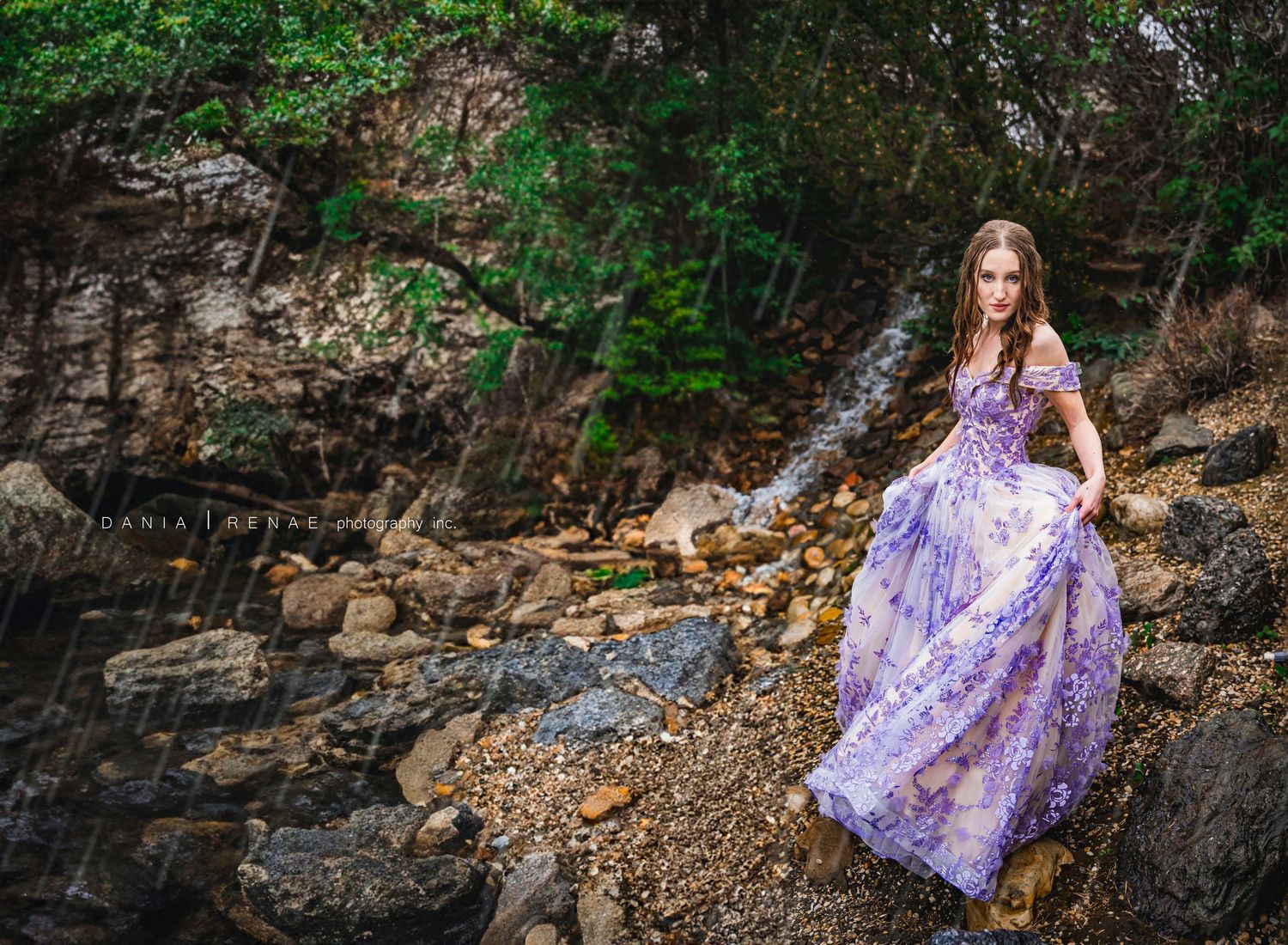 Young woman in elegant purple floral gown posing gracefully on rocky terrain surrounded by lush green trees.