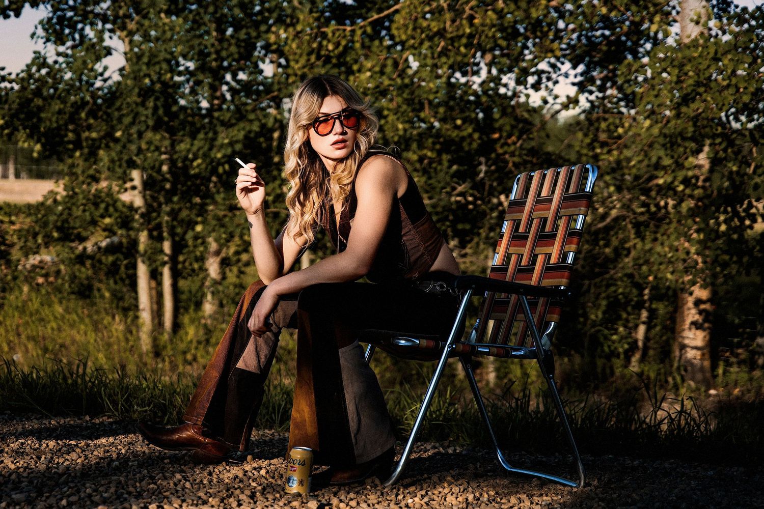Woman in brown jumpsuit with red sunglasses sitting on a folding chair outdoors, holding a cigarette in golden hour light.