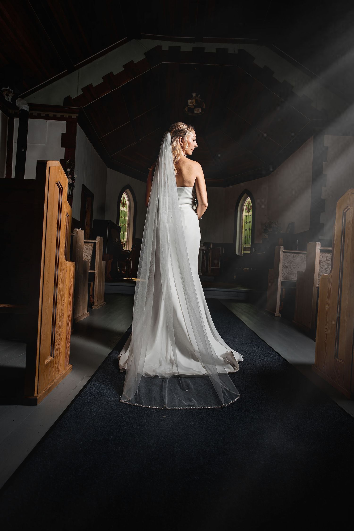 Bride in white gown and long veil stands in sunlit church aisle, viewed from behind.