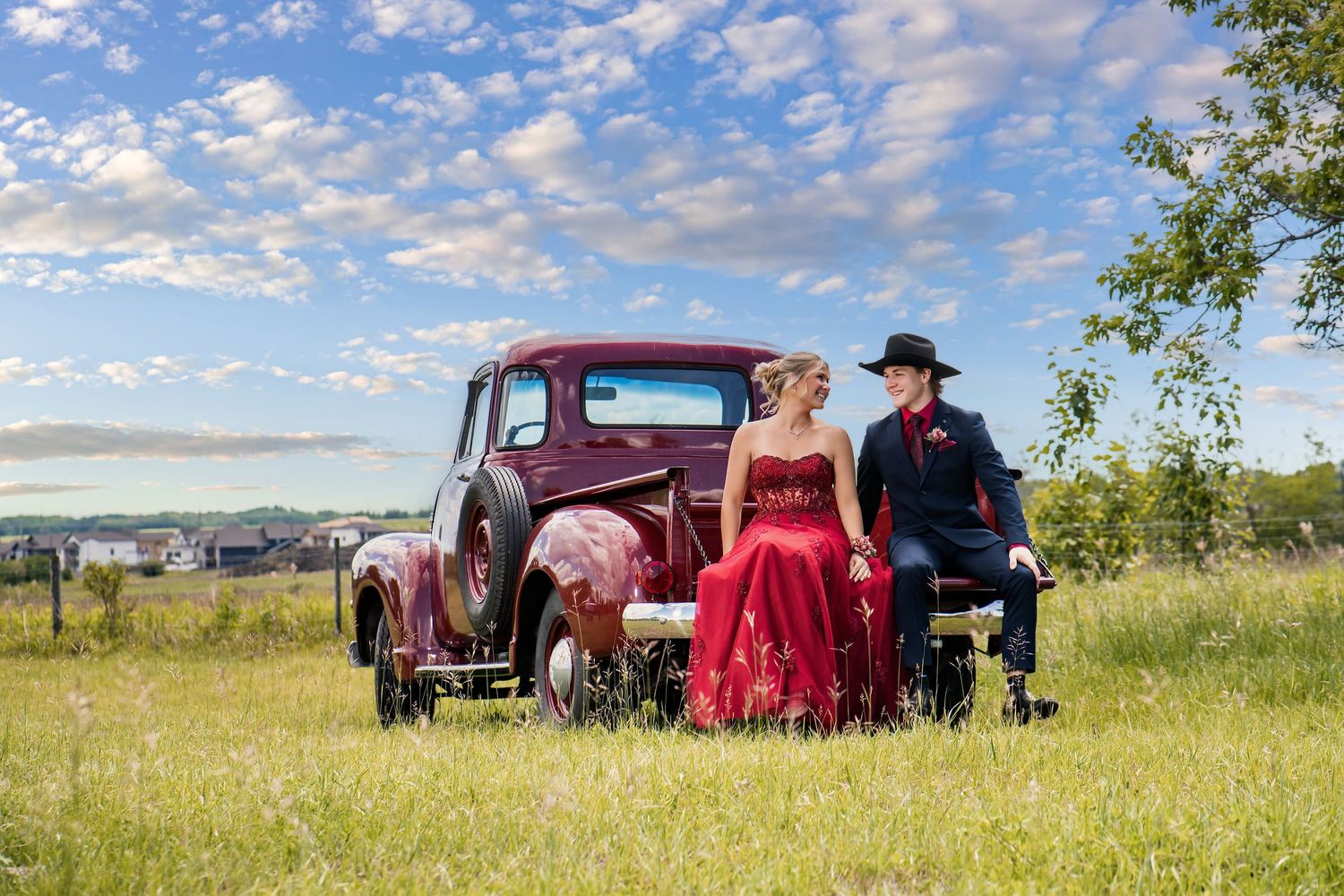 Couple in formal attire posing by a vintage red truck in a green field under a blue sky.