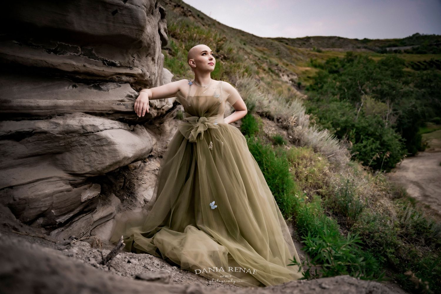 A bald woman in an elegant olive green tulle gown poses gracefully against rocky canyon cliffs with lush greenery.