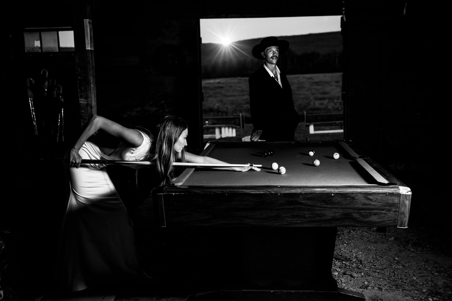 Black and white photo of a woman playing pool indoors while a man stands outside in sunlight.