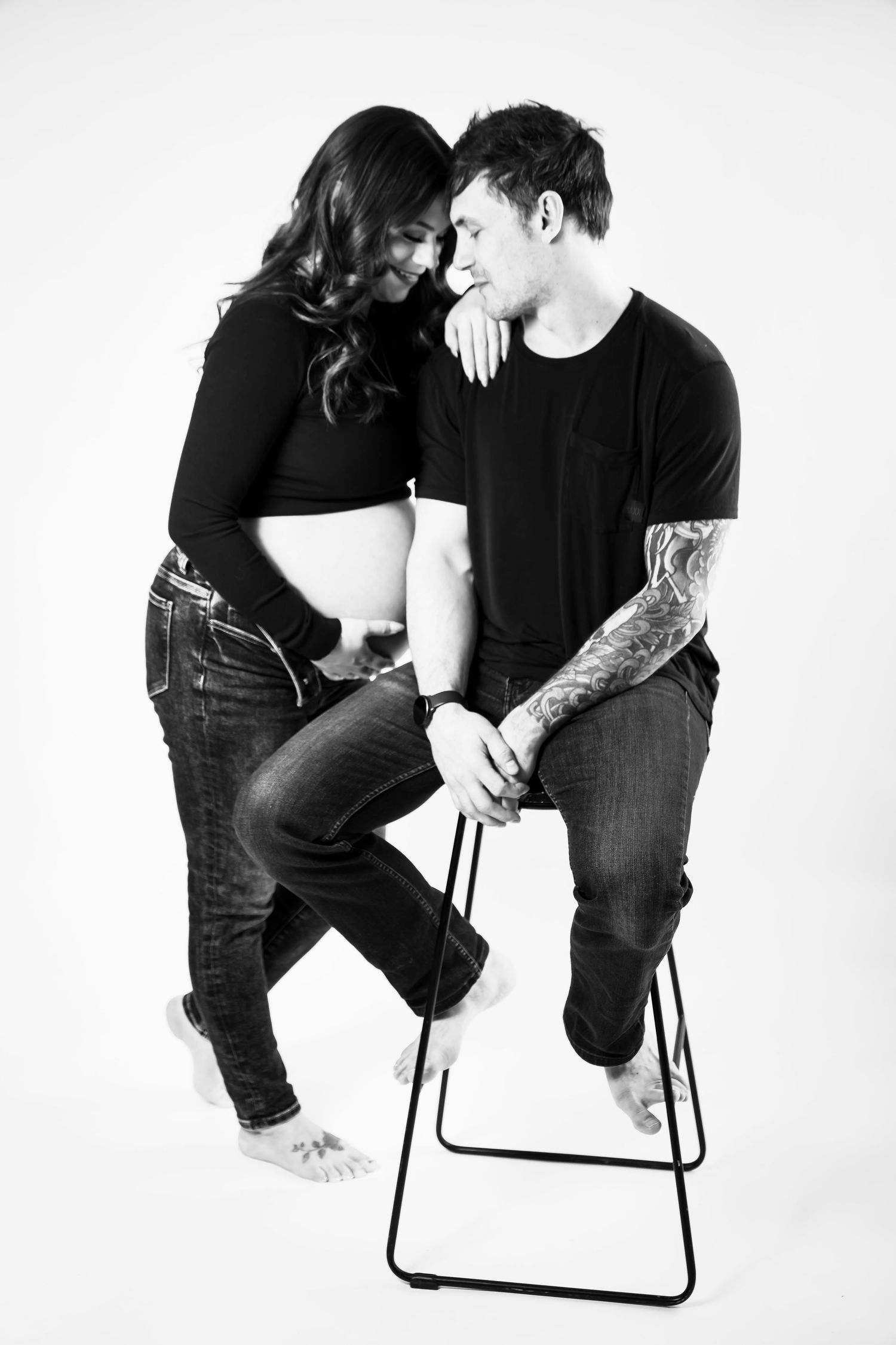 Black and white maternity photo of a couple, man seated on stool with tattoos, woman leaning close showing baby bump.