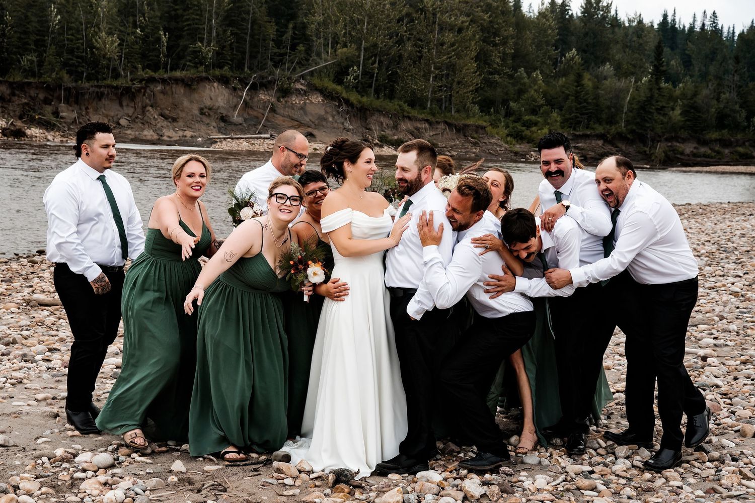 Wedding party laughing on rocky riverbank, bridesmaids in green gowns and groomsmen in white shirts surrounding bride and groom.