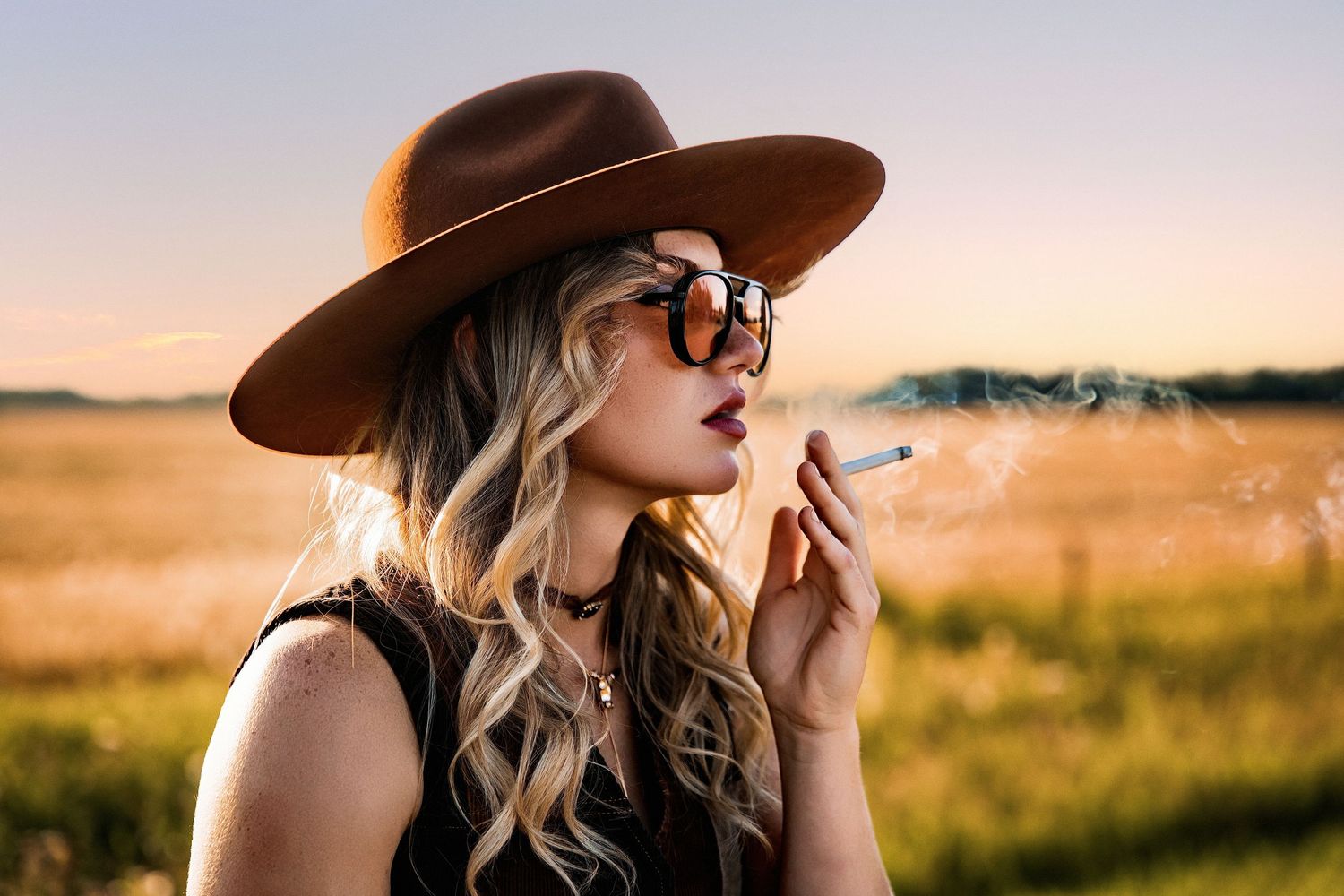 Woman in brown wide-brim hat and sunglasses smoking a cigarette in a golden field at sunset.