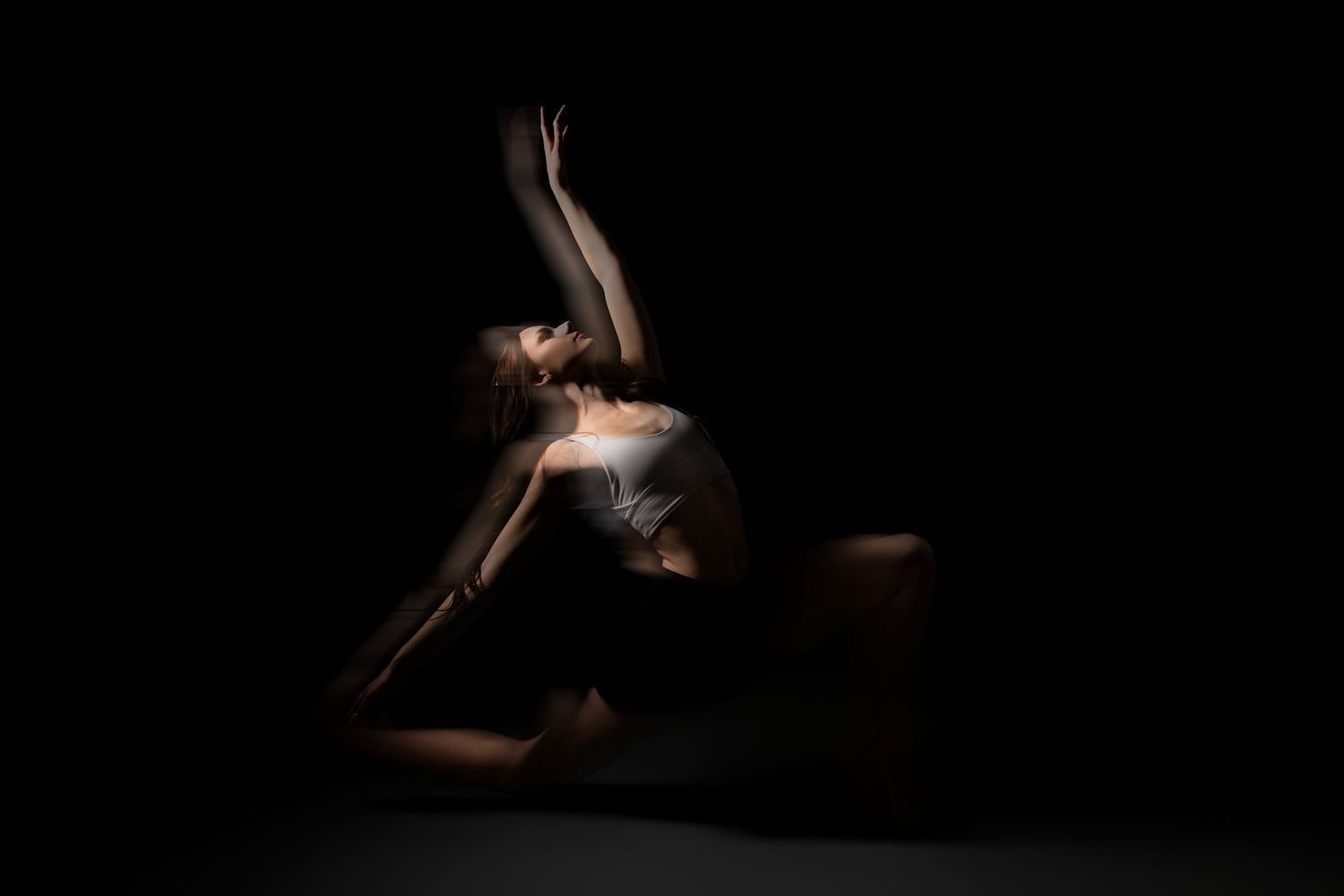 A dancer poses dramatically on the floor in dark studio lighting, arching back with arms raised gracefully.
