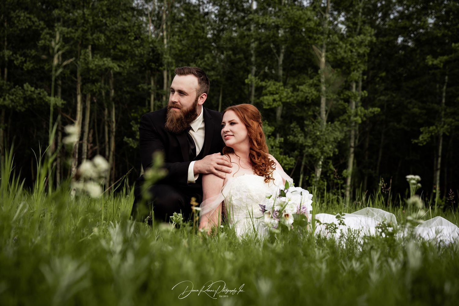 A bearded groom in a dark suit stands behind his bride in a white dress, sitting in a lush green meadow surrounded by trees.