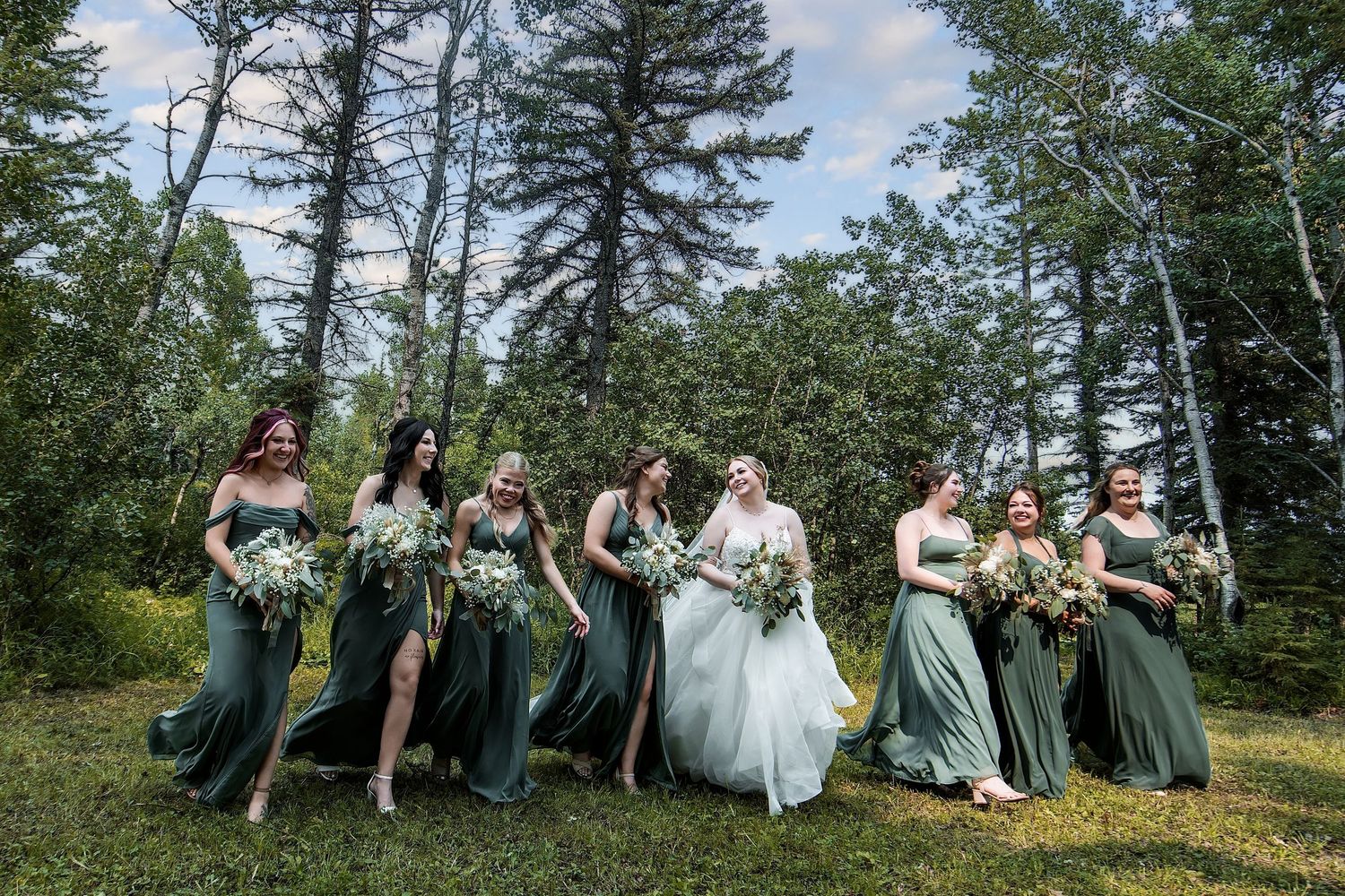 Bride in white gown walks with bridesmaids in green dresses holding bouquets outdoors among tall pine trees.