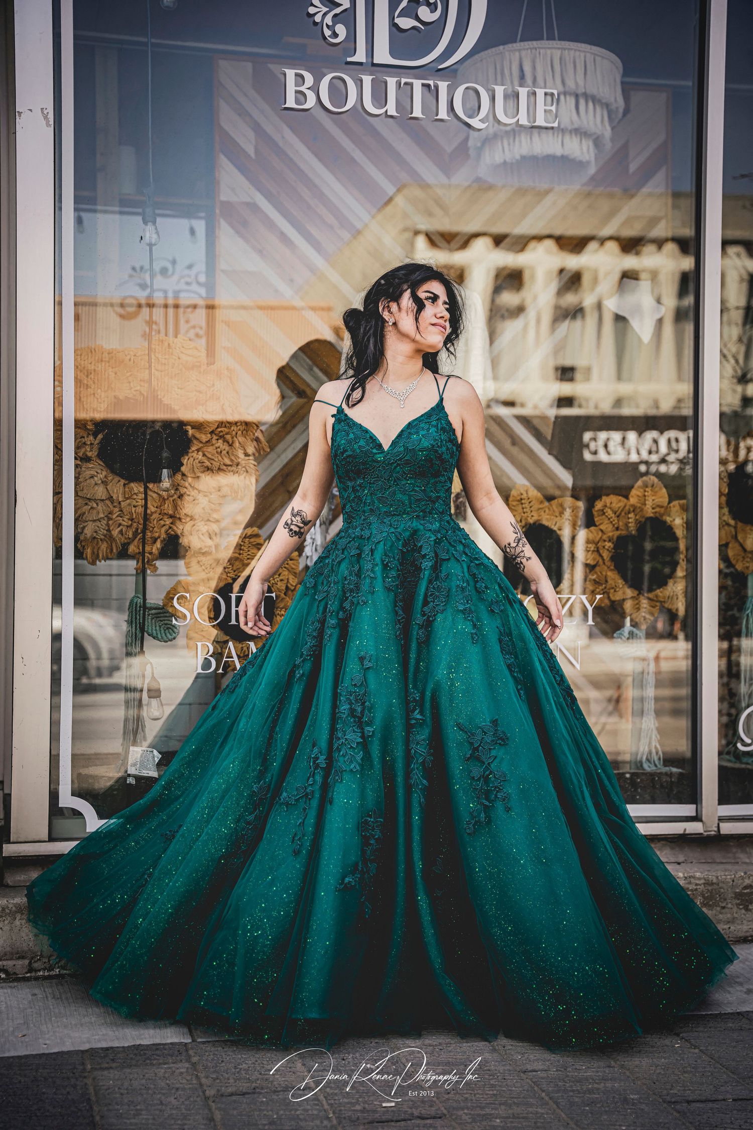 Woman in emerald green ball gown with lace appliques poses elegantly outside a boutique storefront.