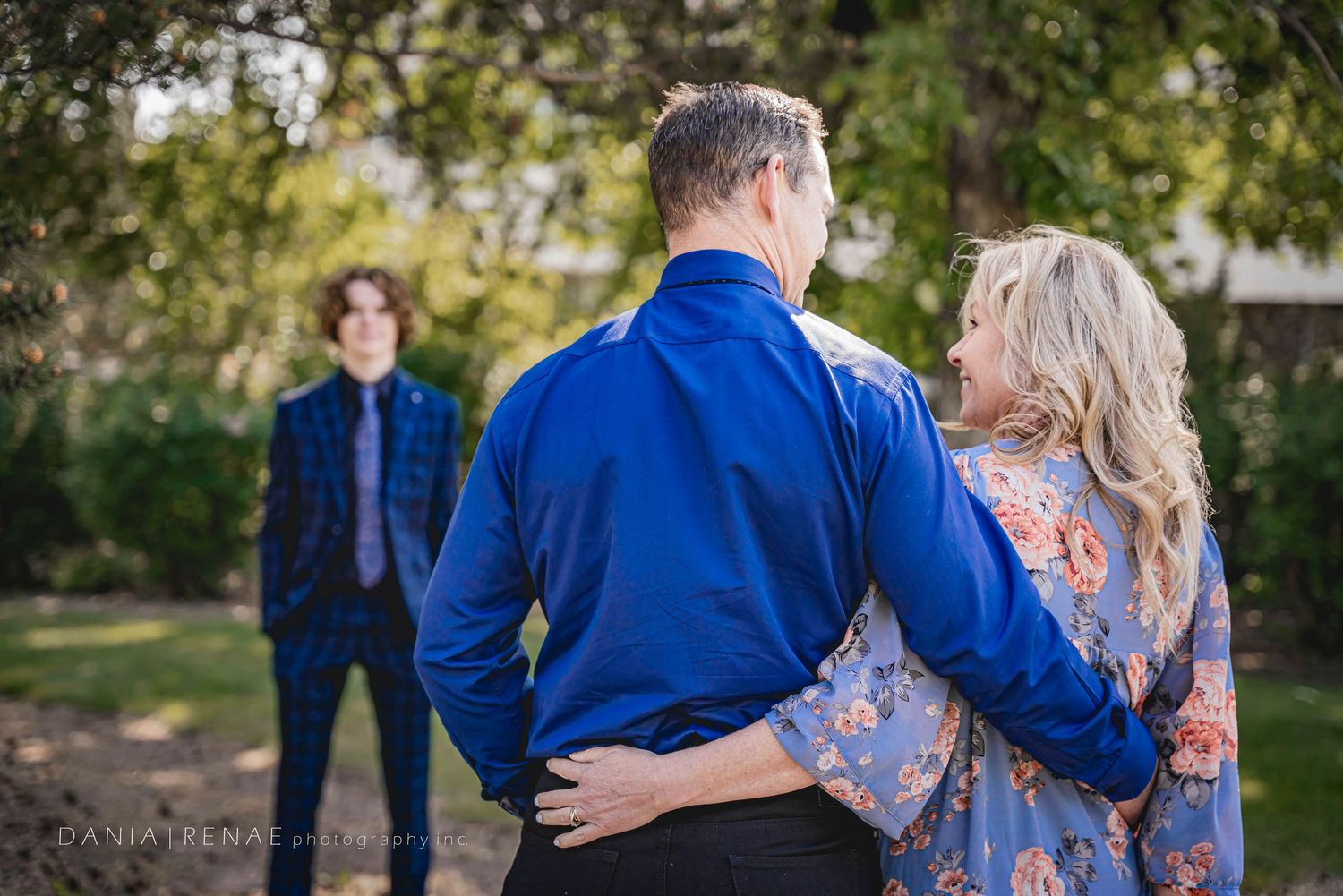 A couple in blue outfits embrace outdoors while a teenage boy stands in the background during a family photo session.