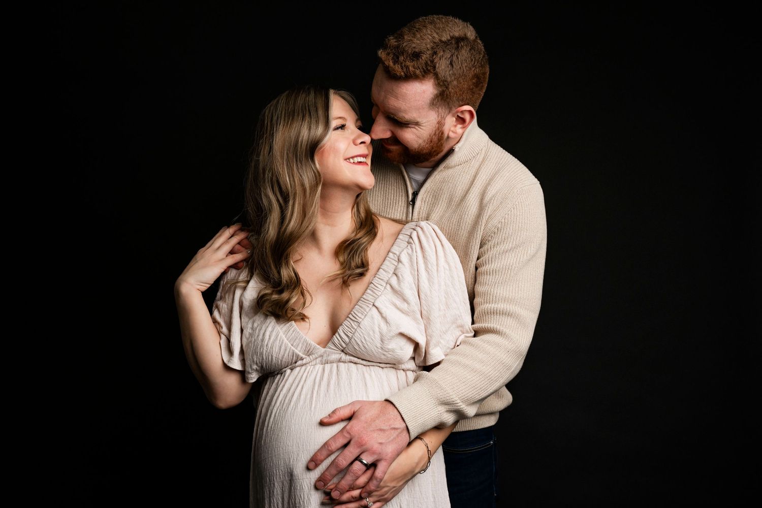 Couple maternity photo shoot, man embracing pregnant woman in flowing dress against dark background.