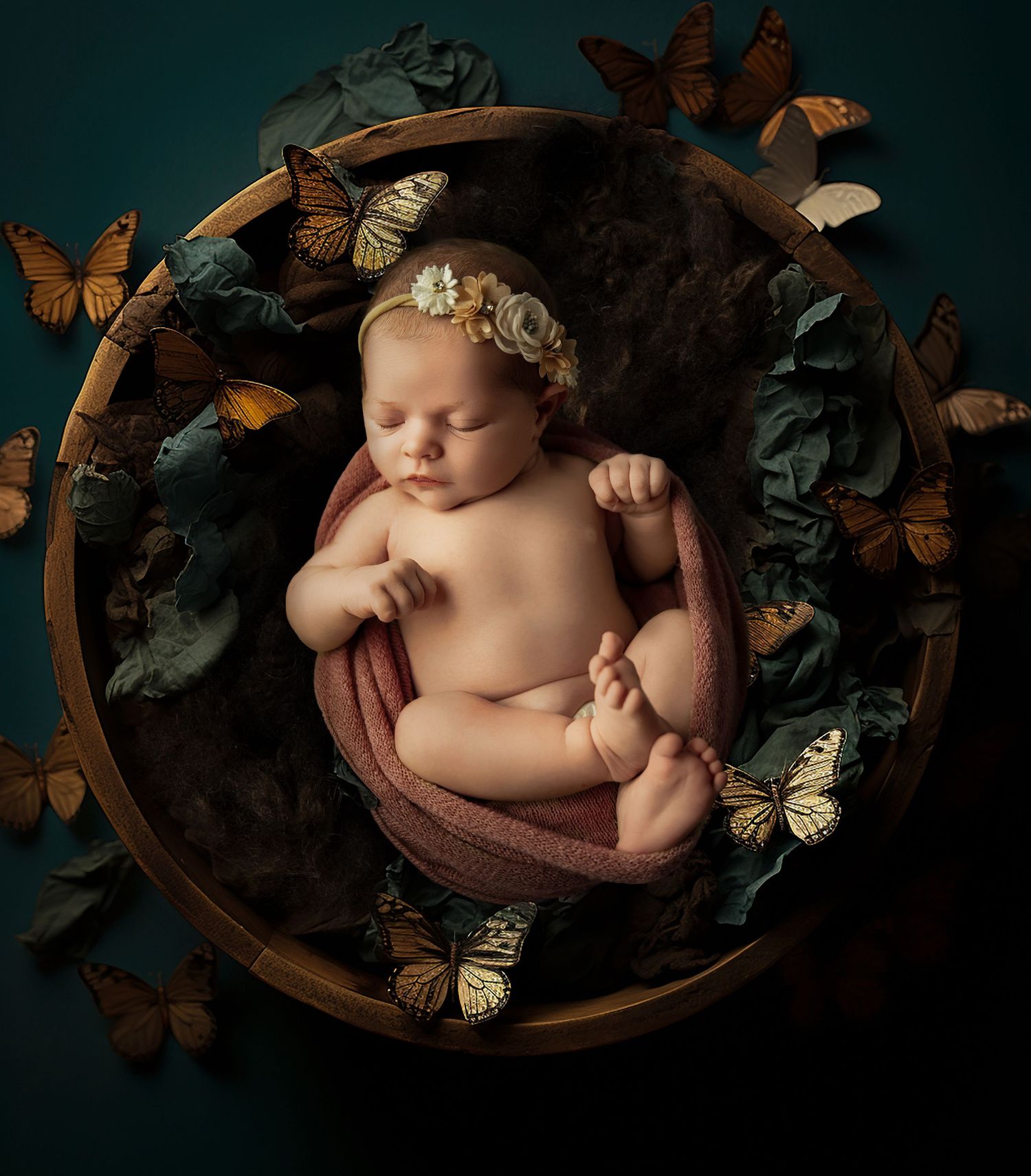 Newborn baby sleeping peacefully in a wooden bowl surrounded by colorful butterflies and teal flowers.