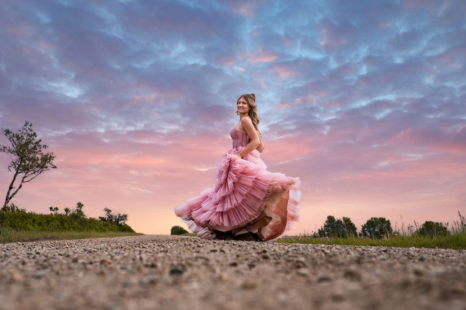 Woman in pink ball gown twirling on road at sunset with dramatic pink and blue cloudy sky background.