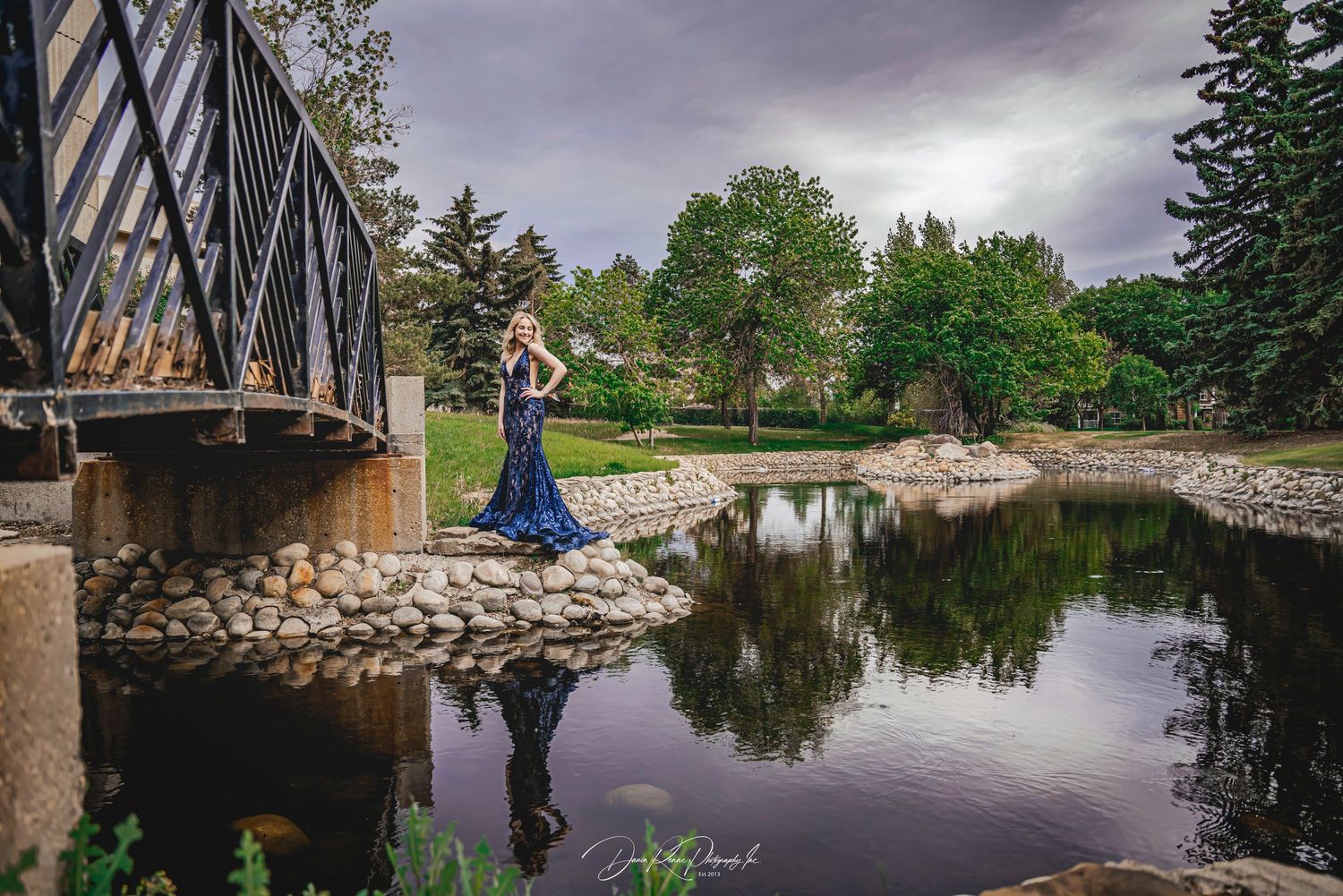 Woman in blue gown poses by a reflective pond near a bridge, surrounded by lush green trees under a cloudy sky.