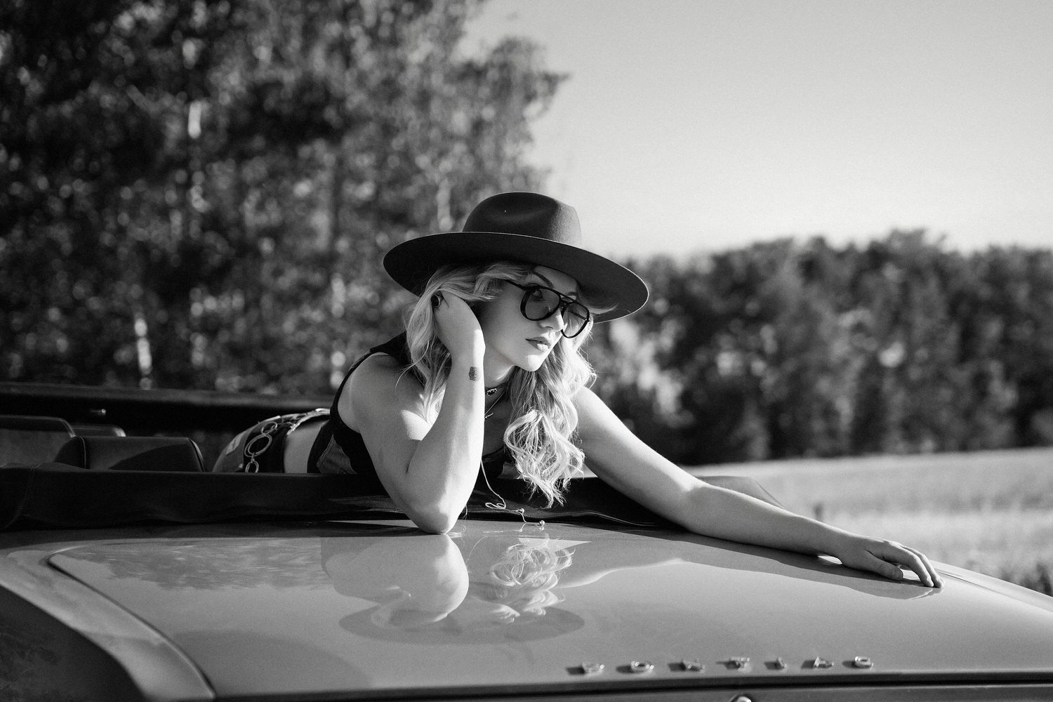 Elegant blonde woman in wide-brim hat and sunglasses posing in a convertible car, black and white photography.