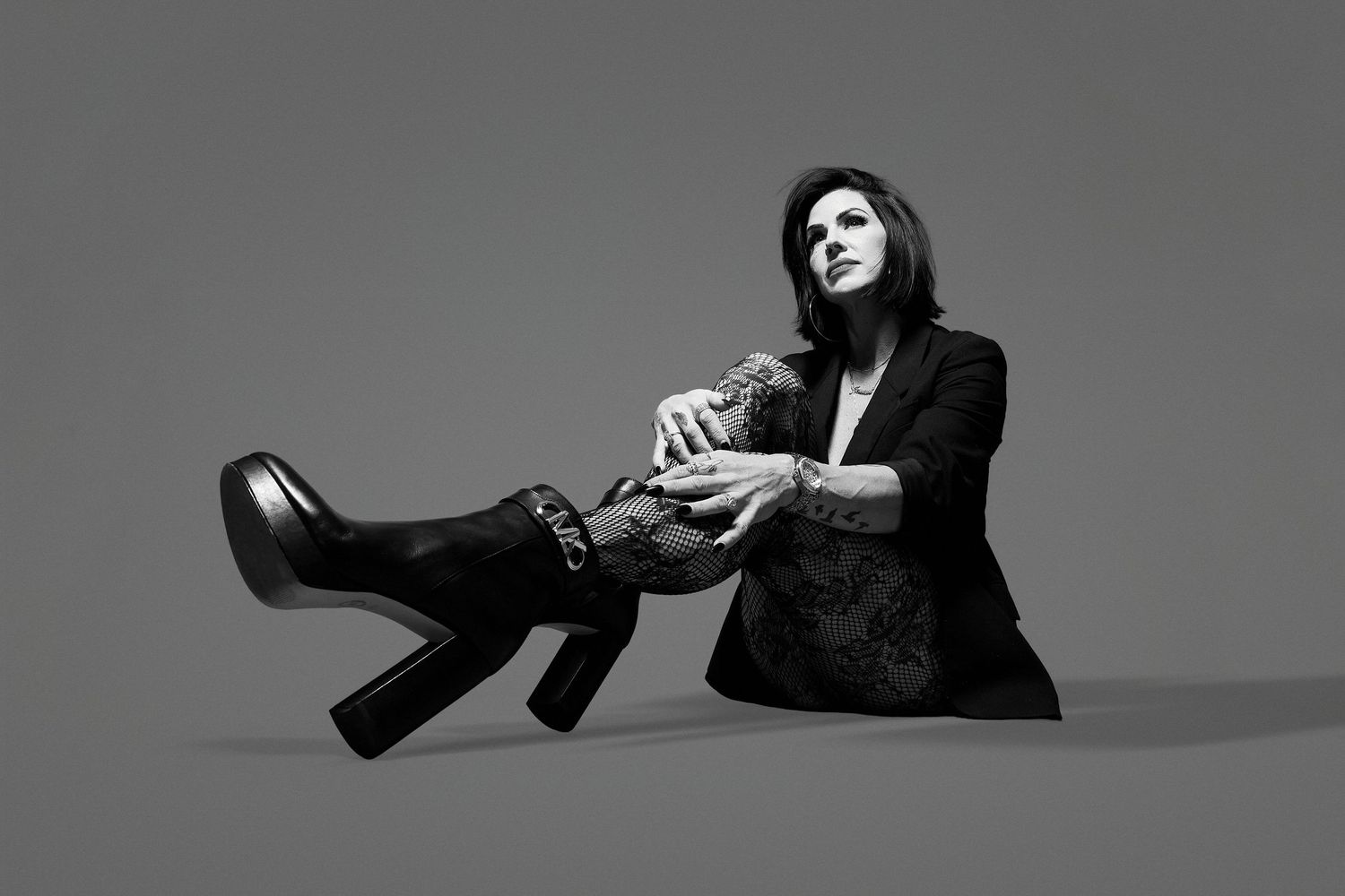 Woman in black blazer and heeled boots seated on floor in dramatic black and white fashion portrait.