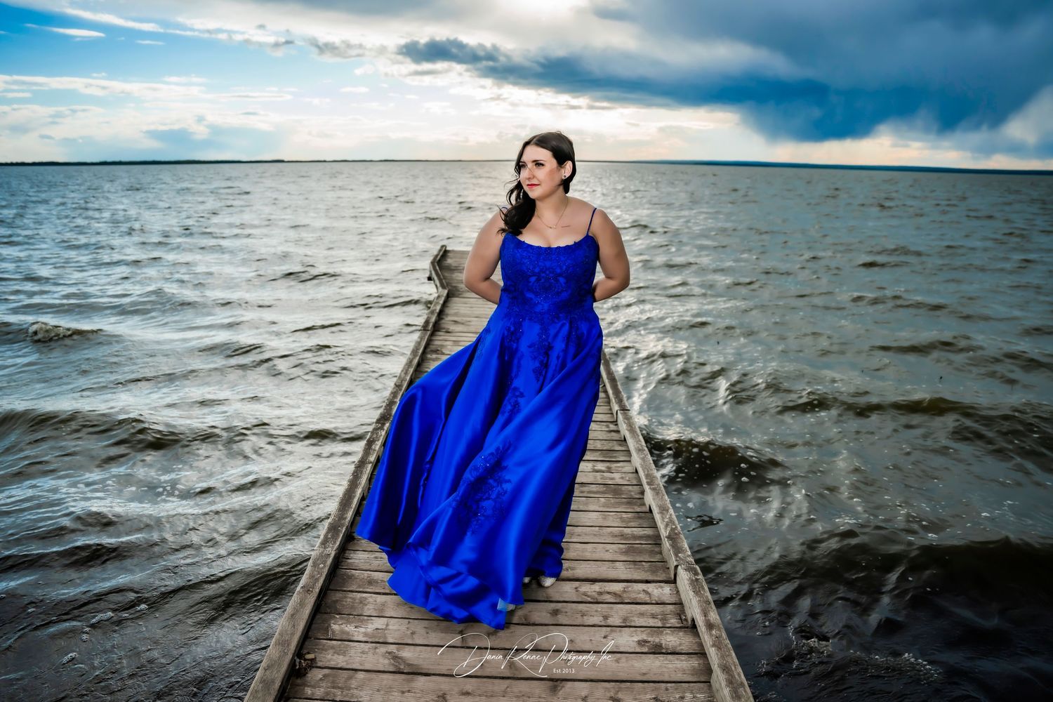 Woman in royal blue gown standing on wooden dock over choppy water under dramatic cloudy sky.