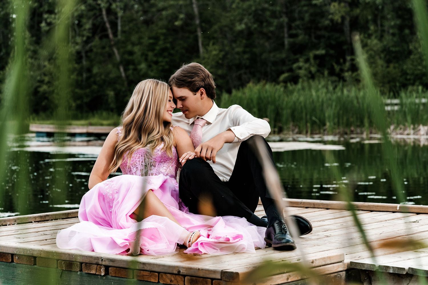 Couple in formal attire sitting on a wooden dock by a serene lake surrounded by lush green trees.
