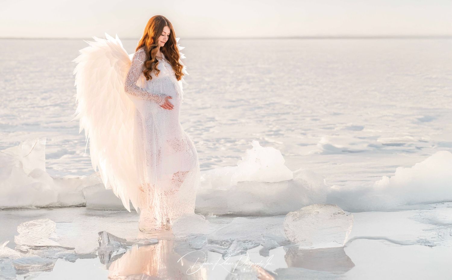 A woman in a white dress with large angel wings stands on a frozen lake surrounded by ice and snow.
