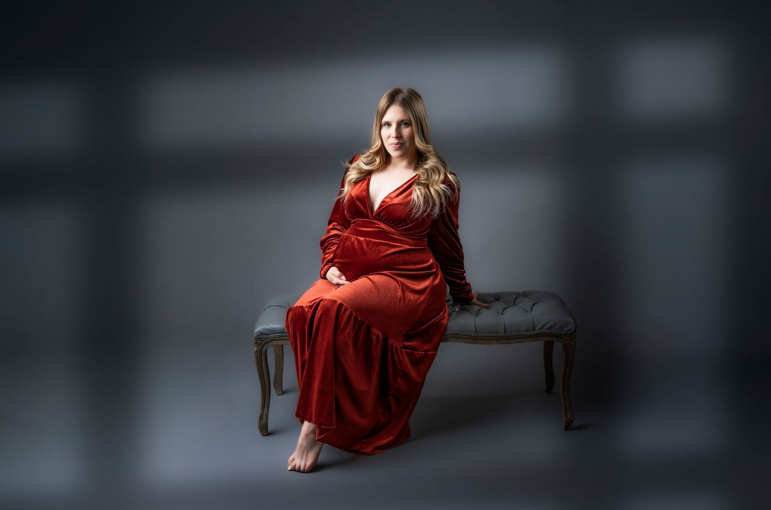 Pregnant woman in elegant red velvet gown seated on tufted bench during professional maternity photo session.