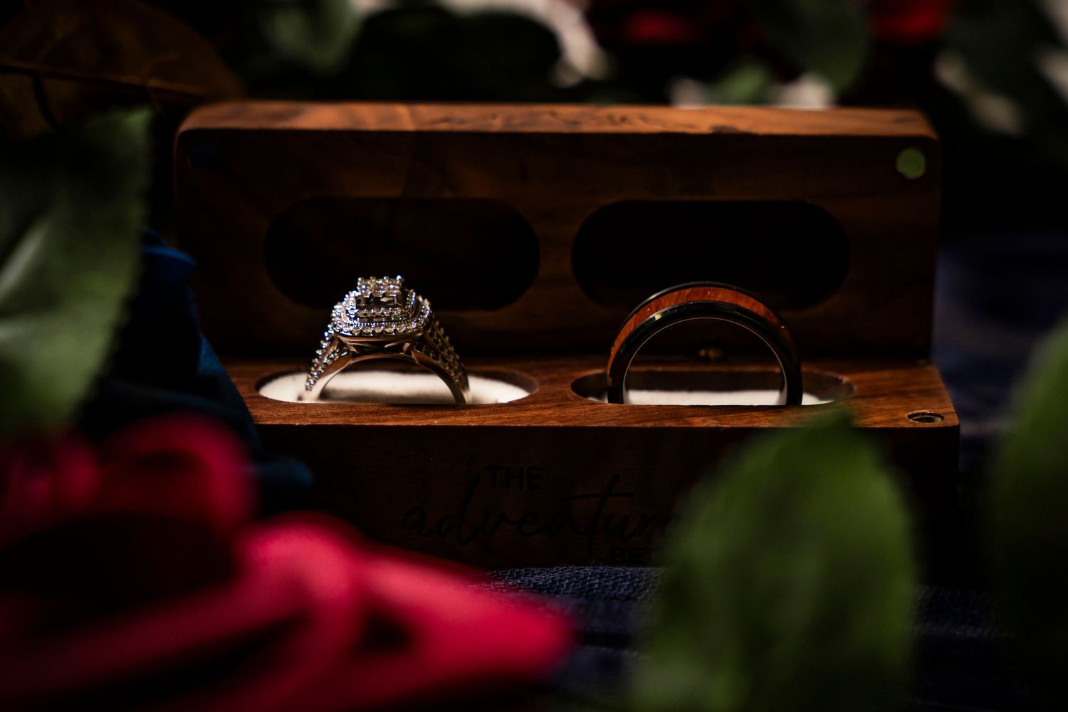 Diamond engagement ring and gold wedding band displayed in a rustic wooden ring box surrounded by red roses.