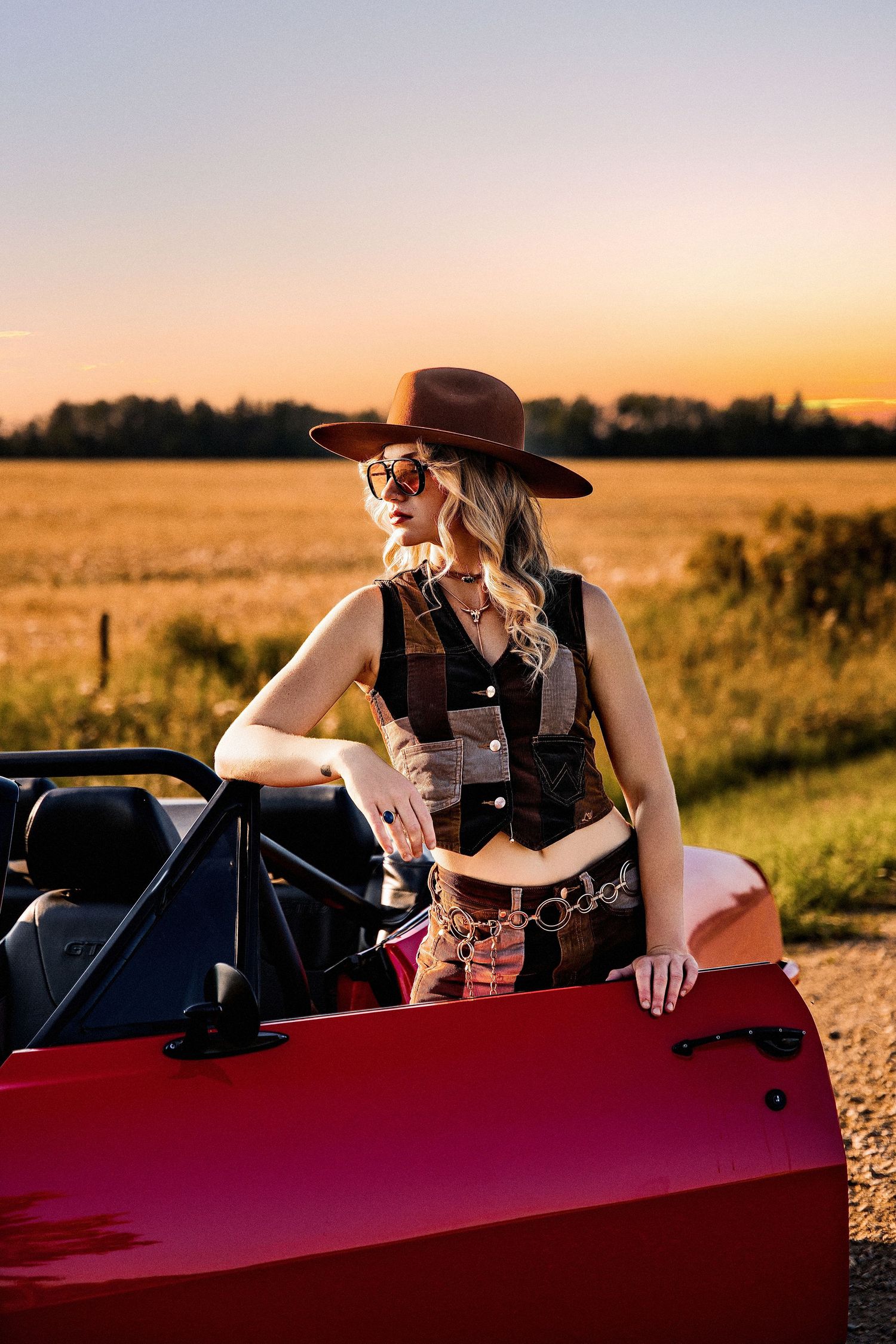 A blonde woman in a cowboy hat and sunglasses leans against a red car in a golden field at sunset.