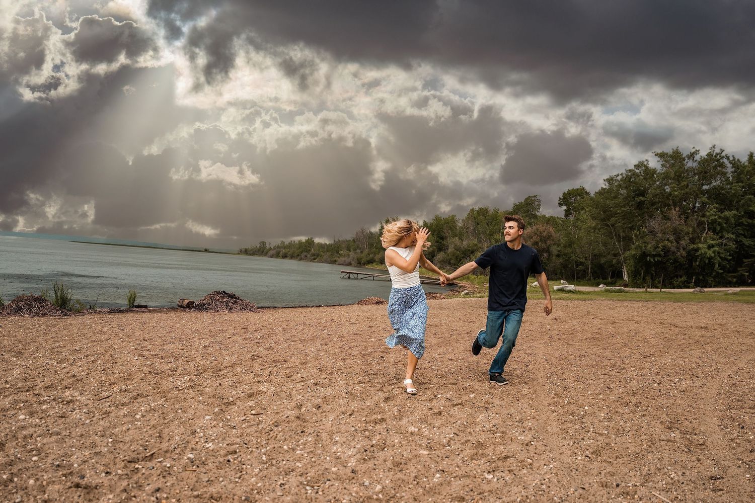 Couple holding hands and running on a pebble beach under dramatic stormy clouds near a calm shoreline.
