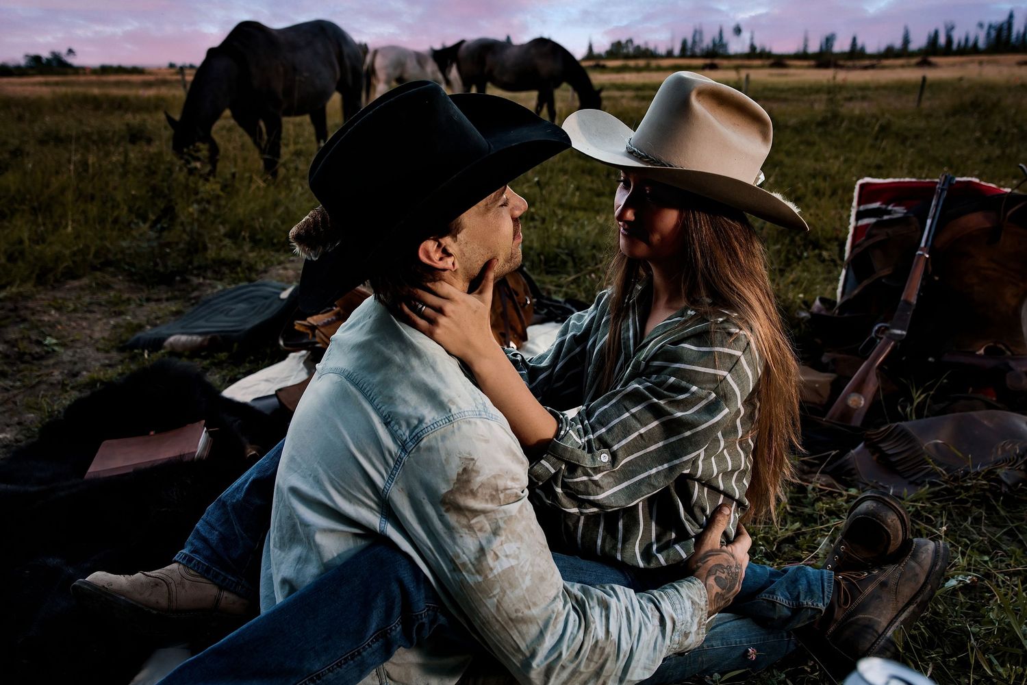 Couple in cowboy hats sharing a romantic moment outdoors at dusk with horses grazing in the background.