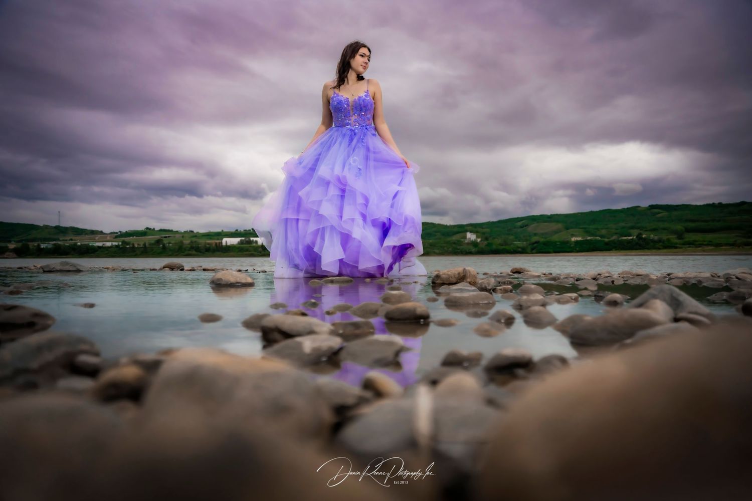 Young woman in flowing purple ball gown standing on rocky riverbank under dramatic cloudy sky.