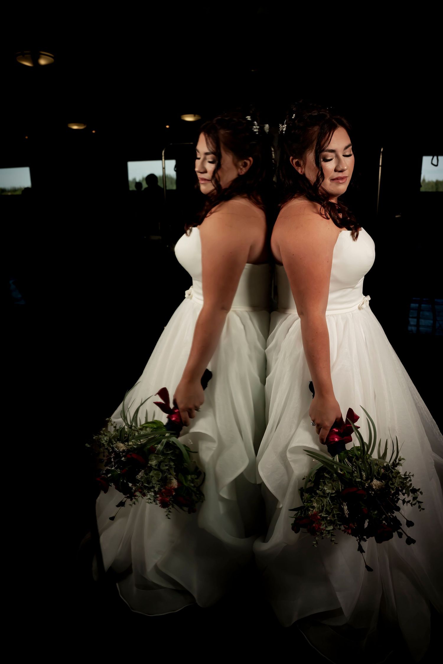 Two brides in white ball gowns holding red floral bouquets stand back-to-back in a dramatic dark setting.