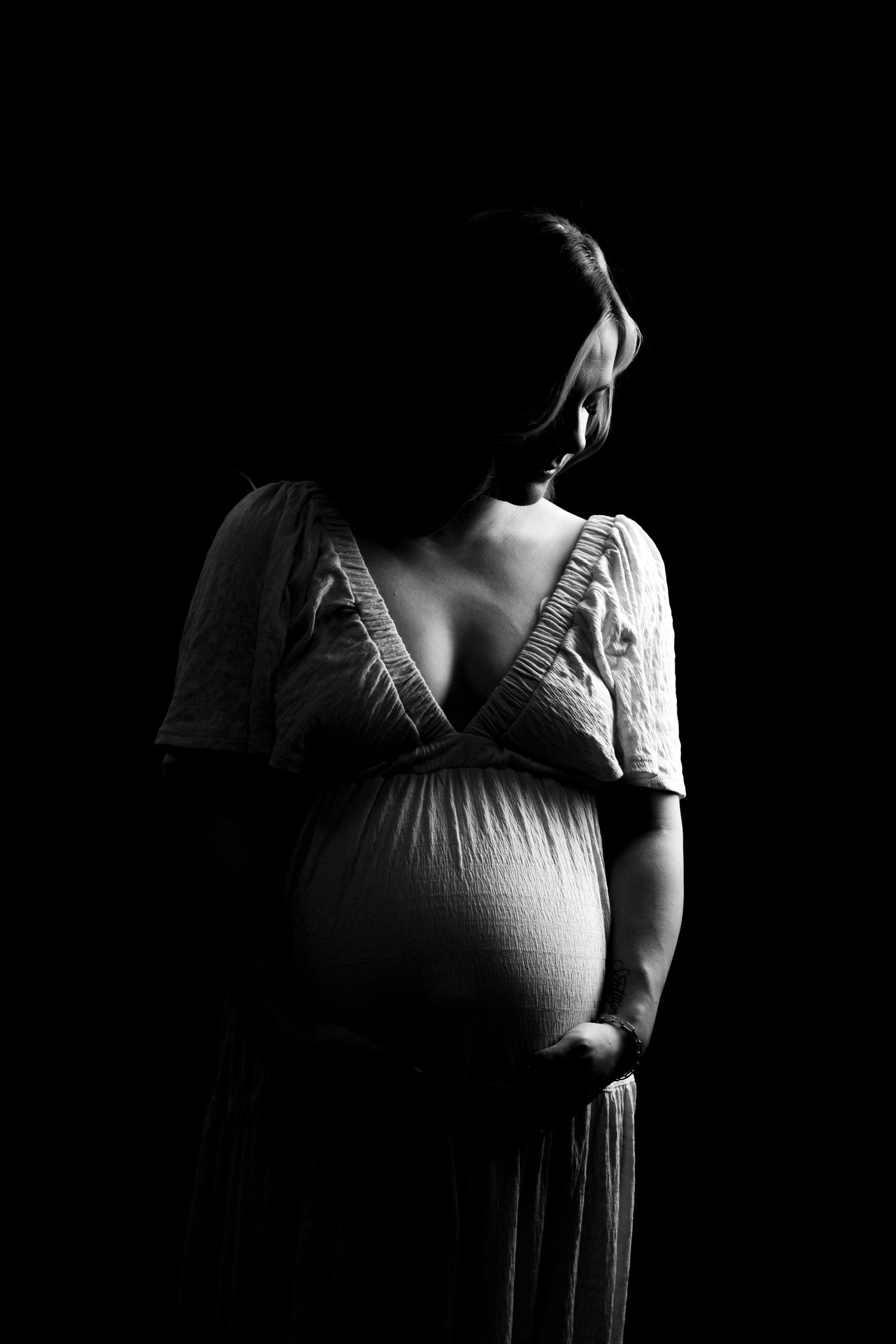 Black and white maternity photo of pregnant woman in white dress cradling baby bump against dark background.