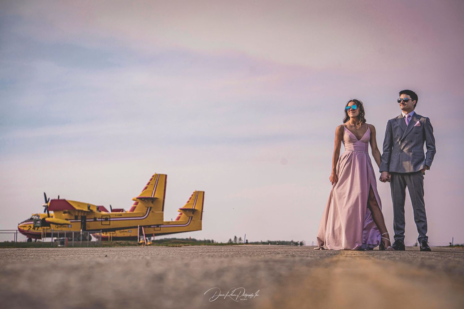 Couple in formal attire holding hands on airstrip with yellow seaplane at sunset.