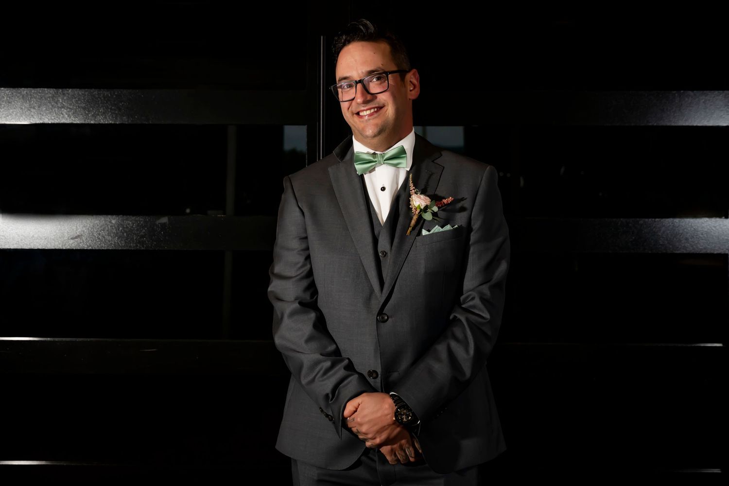 Smiling groom in gray suit with green bow tie and boutonniere poses confidently against dark background.
