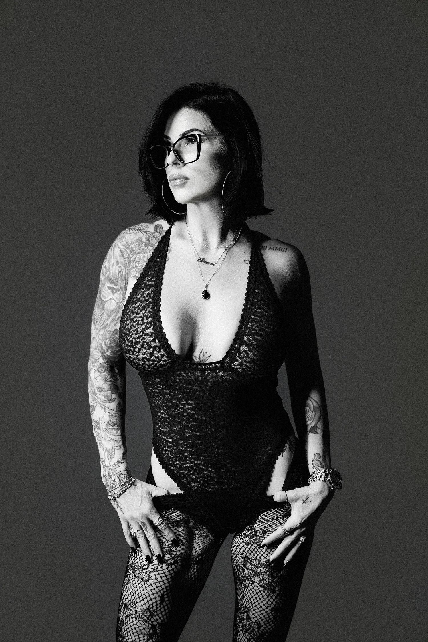 Black and white photo of tattooed woman in lace bodysuit and fishnet tights wearing oversized glasses posing in studio.