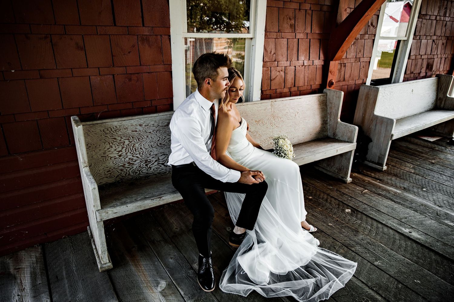 A bride and groom share an intimate moment on a rustic wooden bench outside a red barn on their wedding day.